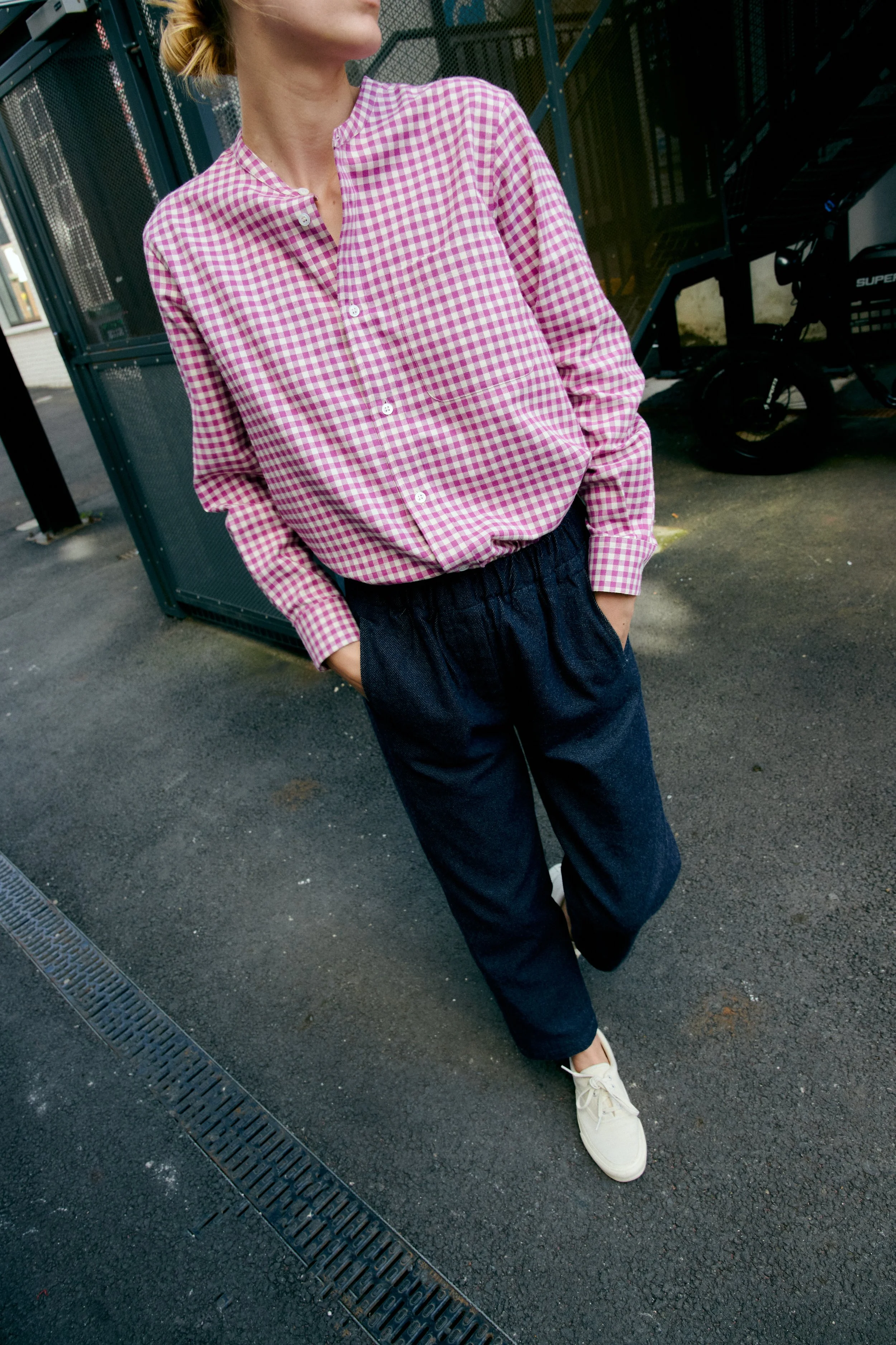Person wearing a pink checkered shirt, dark pants, and white shoes standing outdoors near a metal grate and a staircase.
