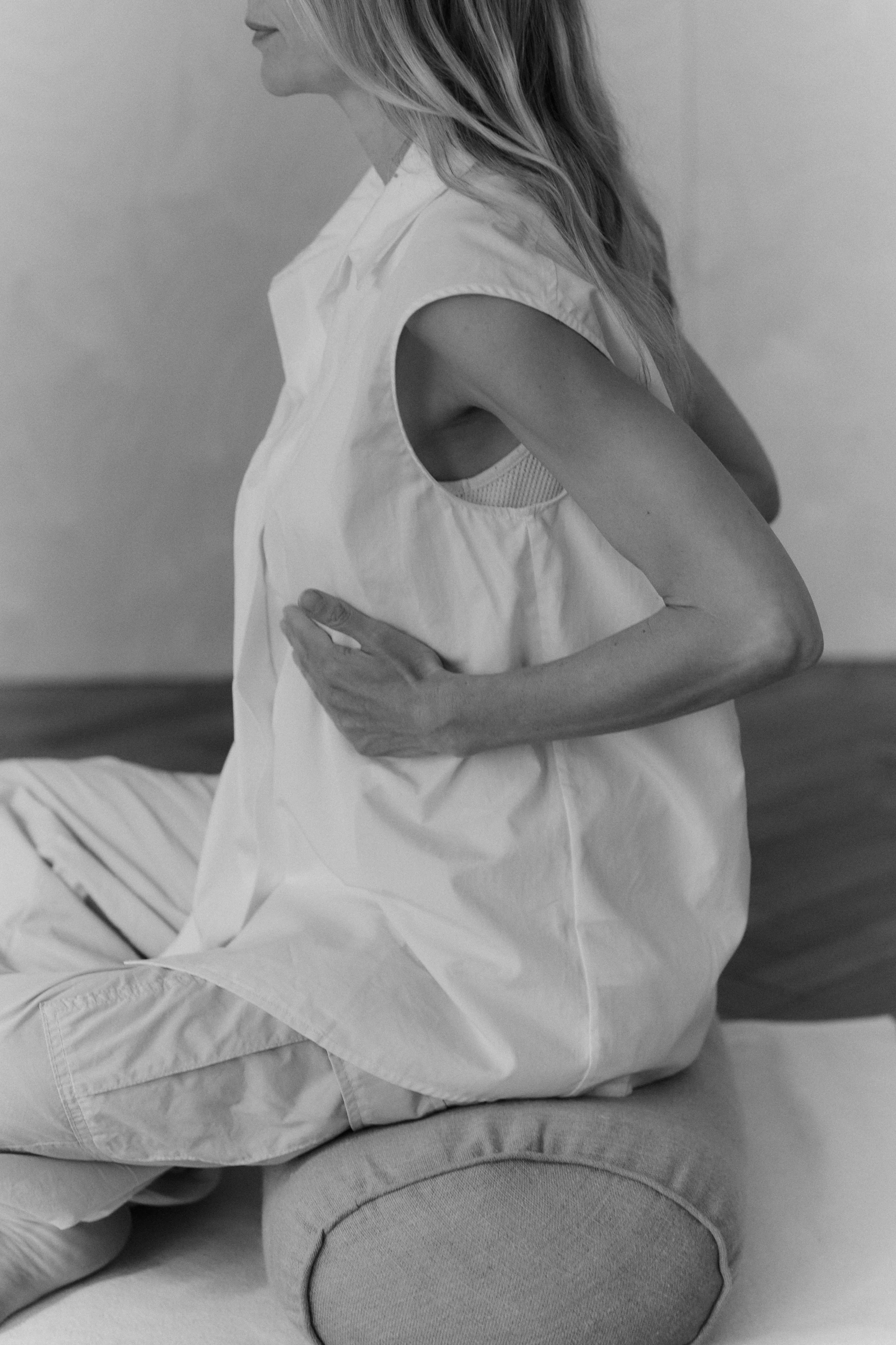 Black and white photo of a woman sitting cross-legged on a yoga mat, wearing a loose shirt and pants, with her hand on her abdomen.