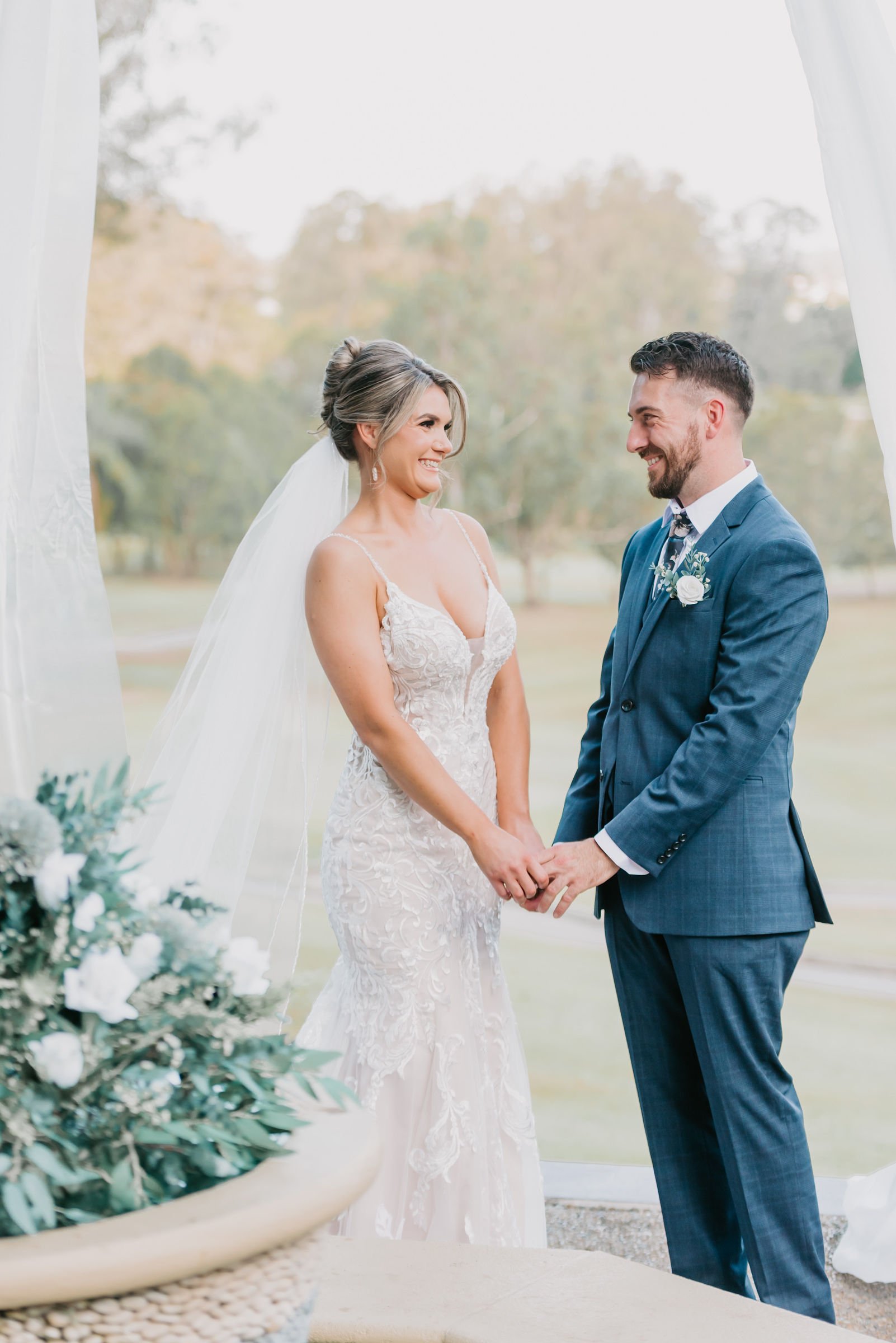 Bride and groom holding hands during wedding ceremony outside, smiling at each other, with trees in the background.