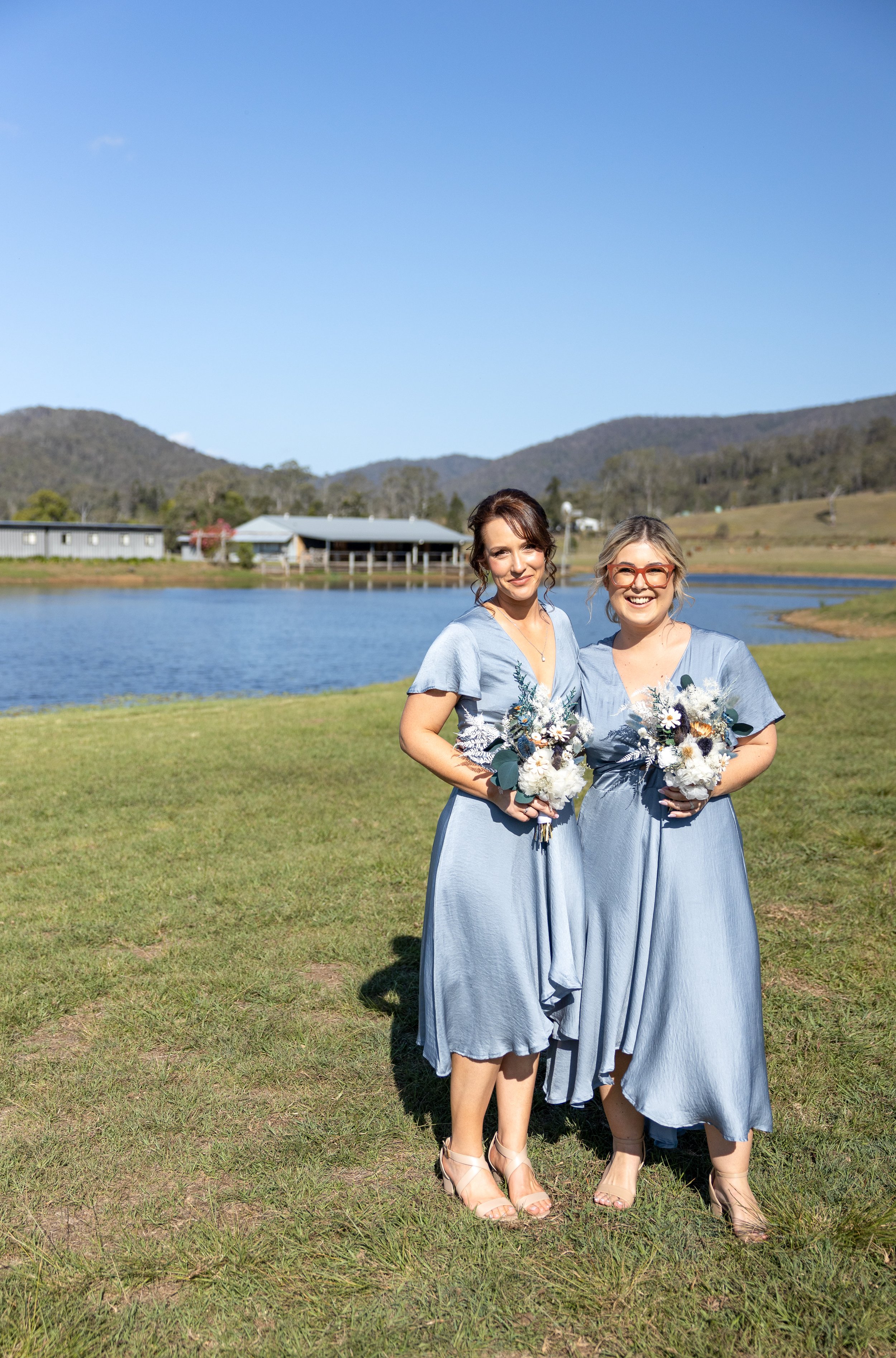Two women in matching light blue dresses holding bouquets, standing on grass near a lake with rolling hills in the background on a sunny day.