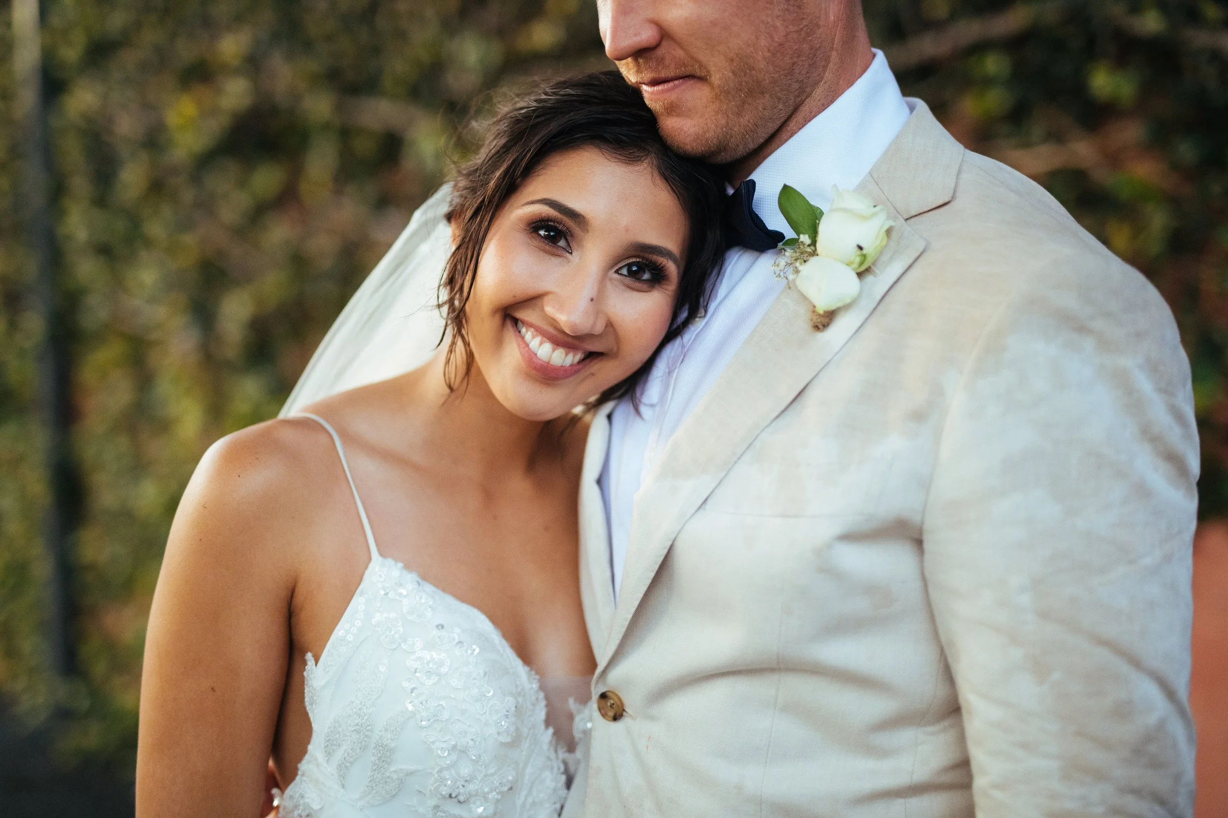 A bride smiling and leaning her head on a groom's chest during a wedding, with trees in the background.