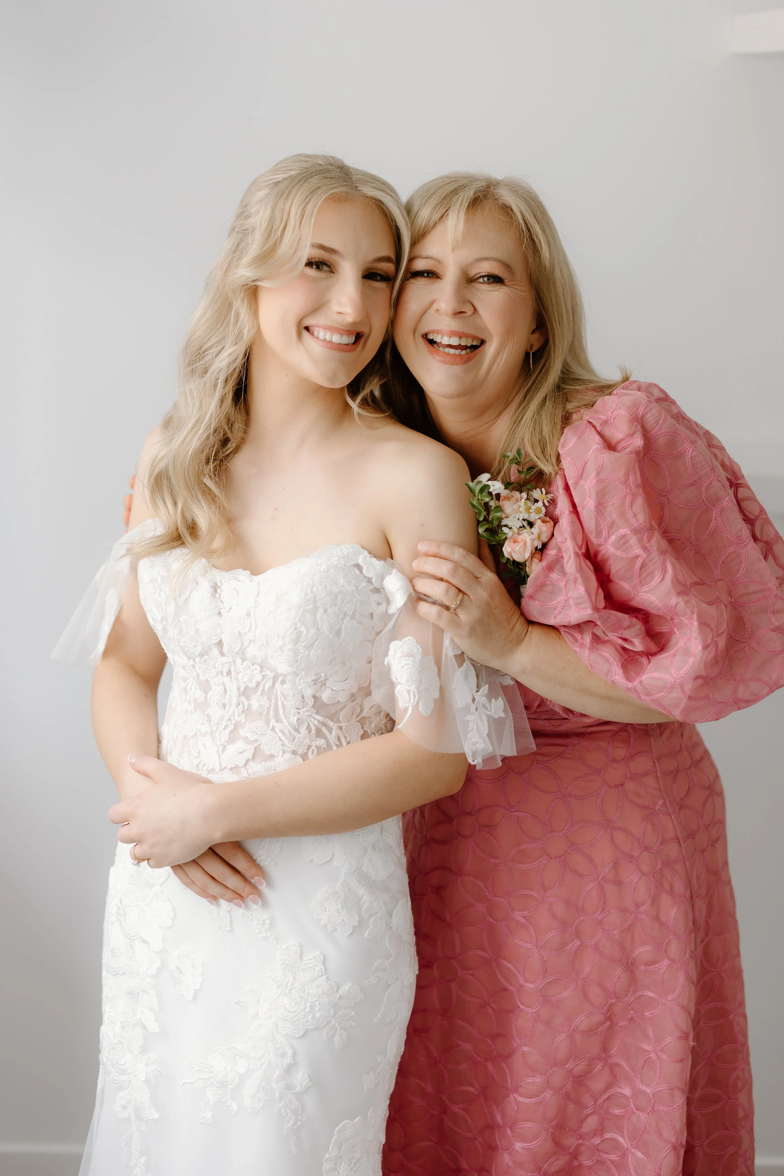 A young woman in a white lace wedding dress smiling with an older woman in a pink dress, both embracing and smiling, against a plain light background.