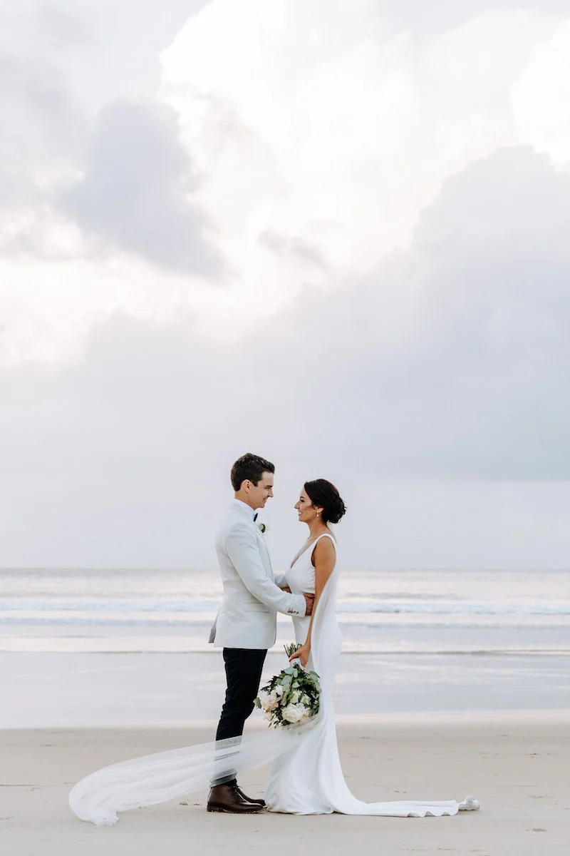 A bride and groom standing on the beach, holding hands, with the ocean and cloudy sky in the background.