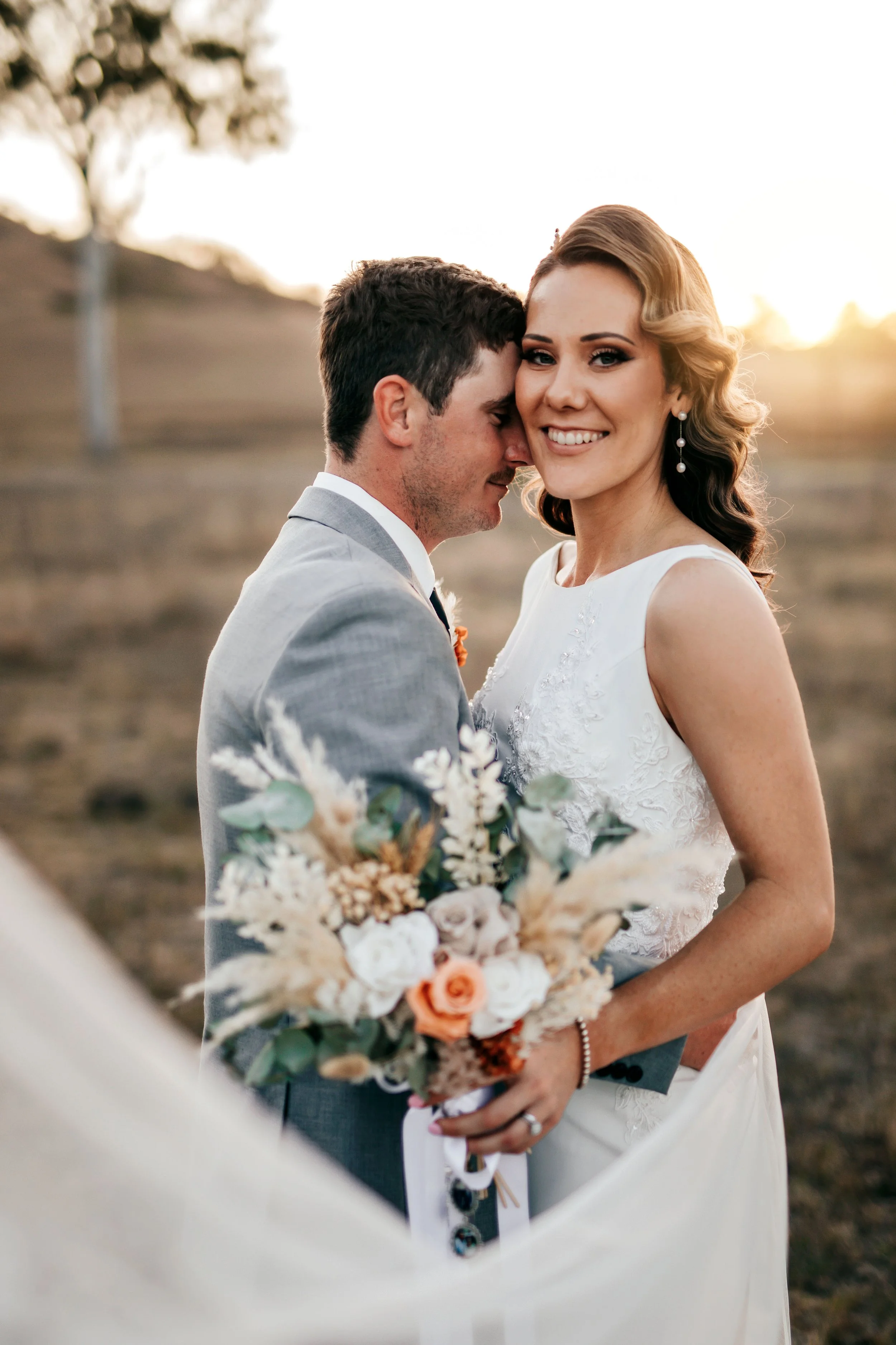 A newlywed couple standing outdoors at sunset, with the groom hugging the bride and resting his forehead on her temple, both smiling. The bride is holding a bouquet of flowers, and the background features a field with a tree.