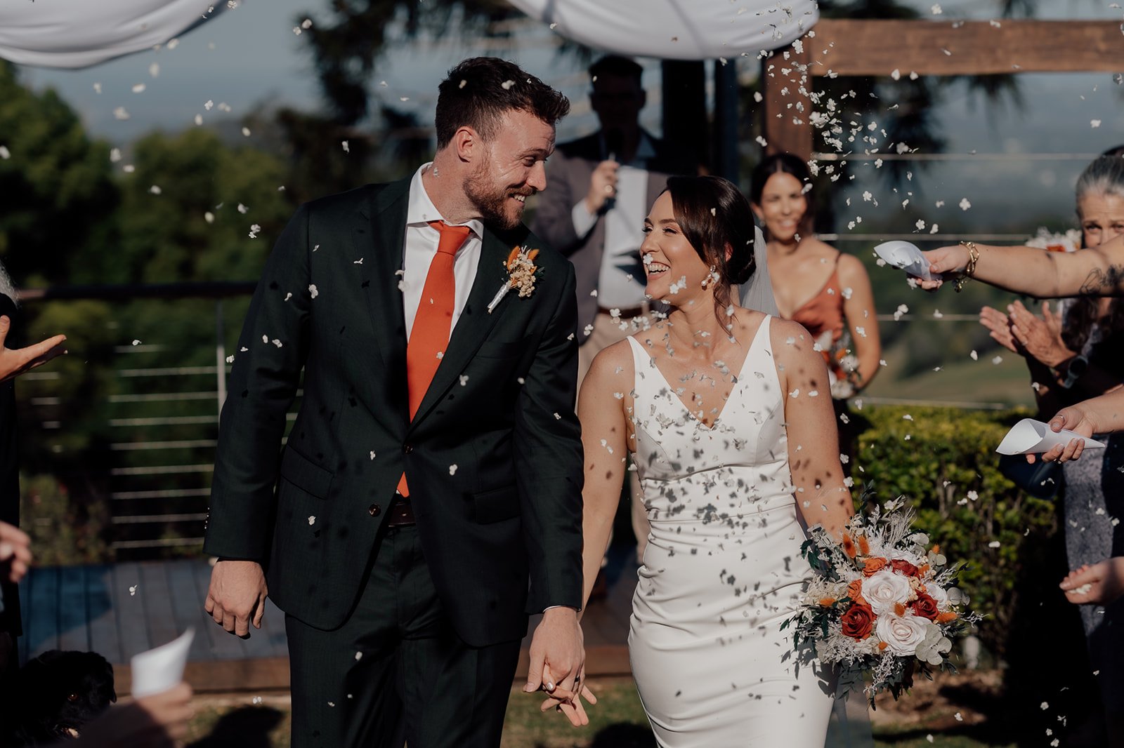A newlywed couple walking hand-in-hand amidst confetti at their outdoor wedding ceremony, smiling and surrounded by friends and family.