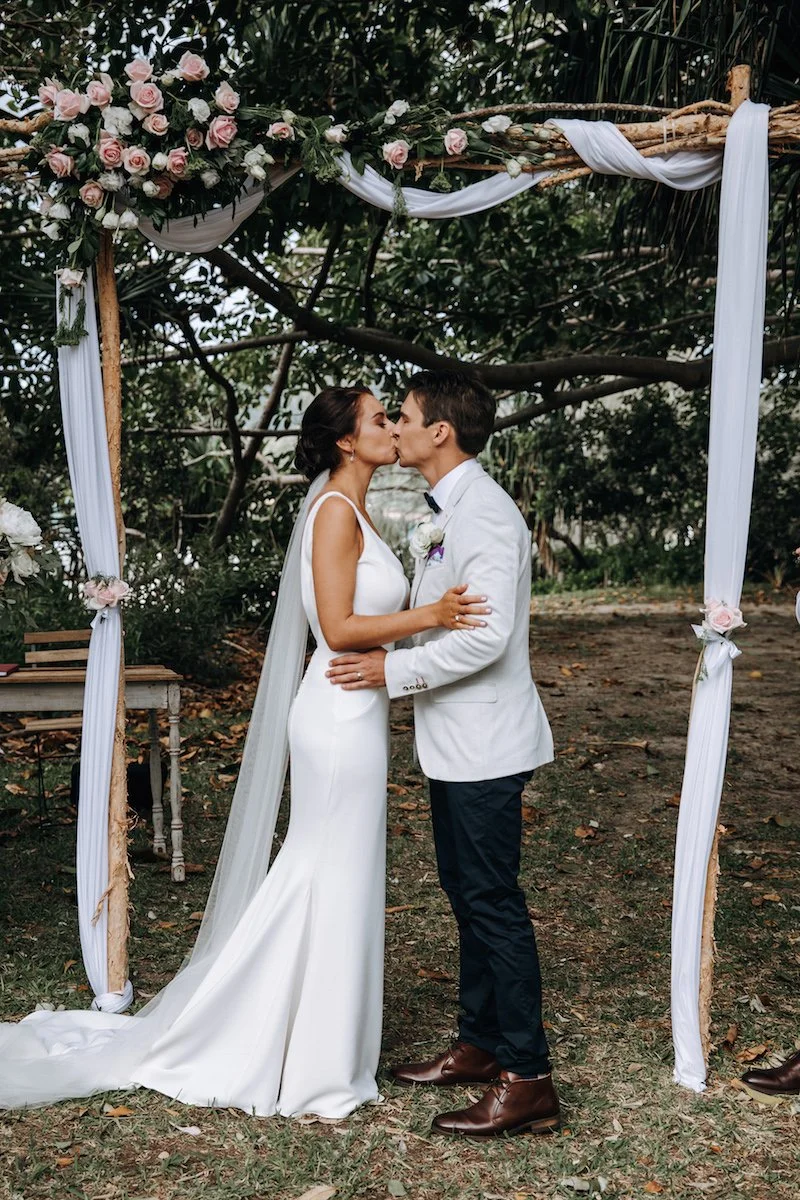 A bride and groom kiss under a wooden wedding arch decorated with pink and white roses and white fabric, outdoors in a natural setting.