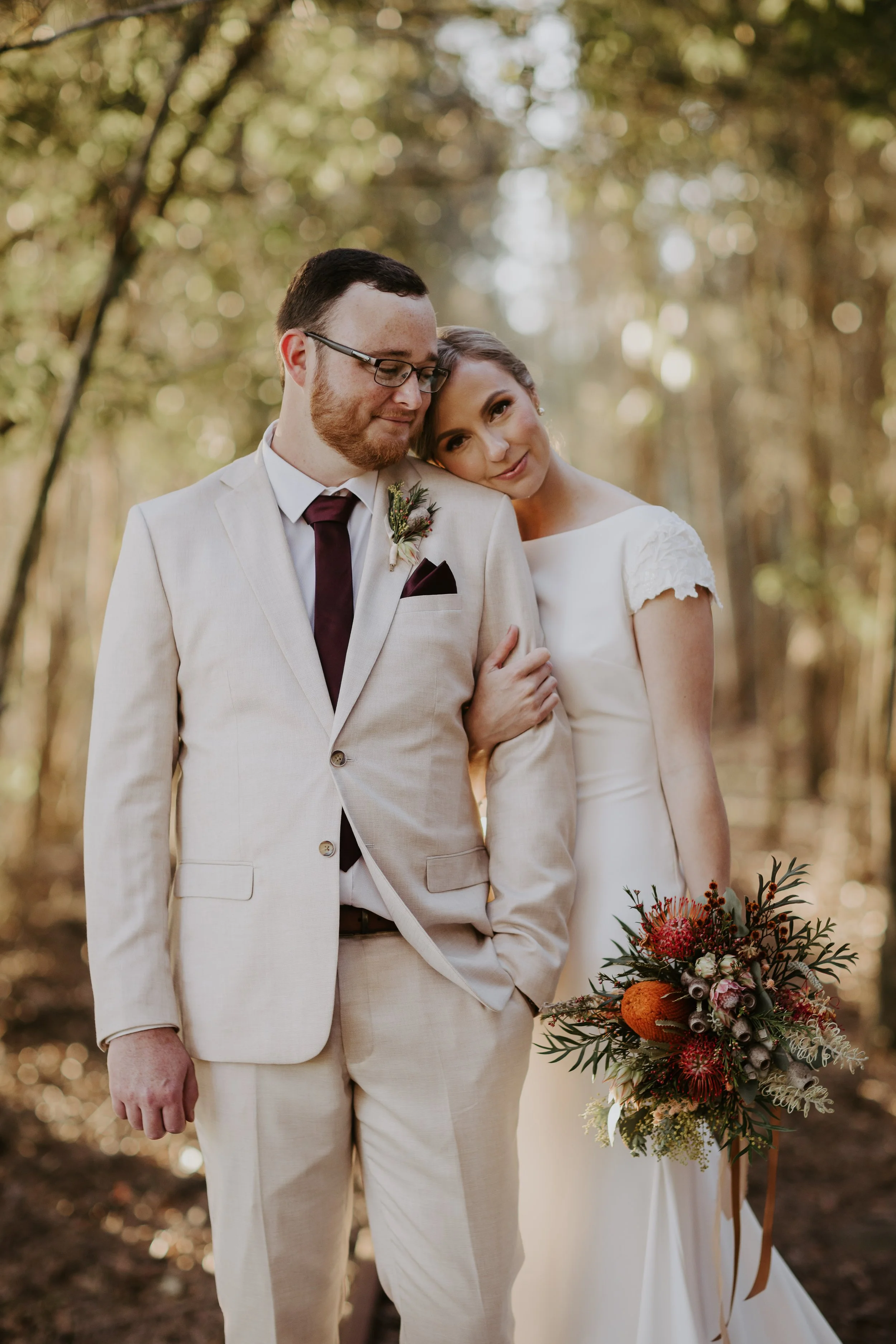 A bride and groom stand closely together in a wooded outdoor setting, with the bride resting her head on the groom's shoulder. The groom wears a light-colored suit with a dark tie and glasses, while the bride wears a white dress with lace sleeves and