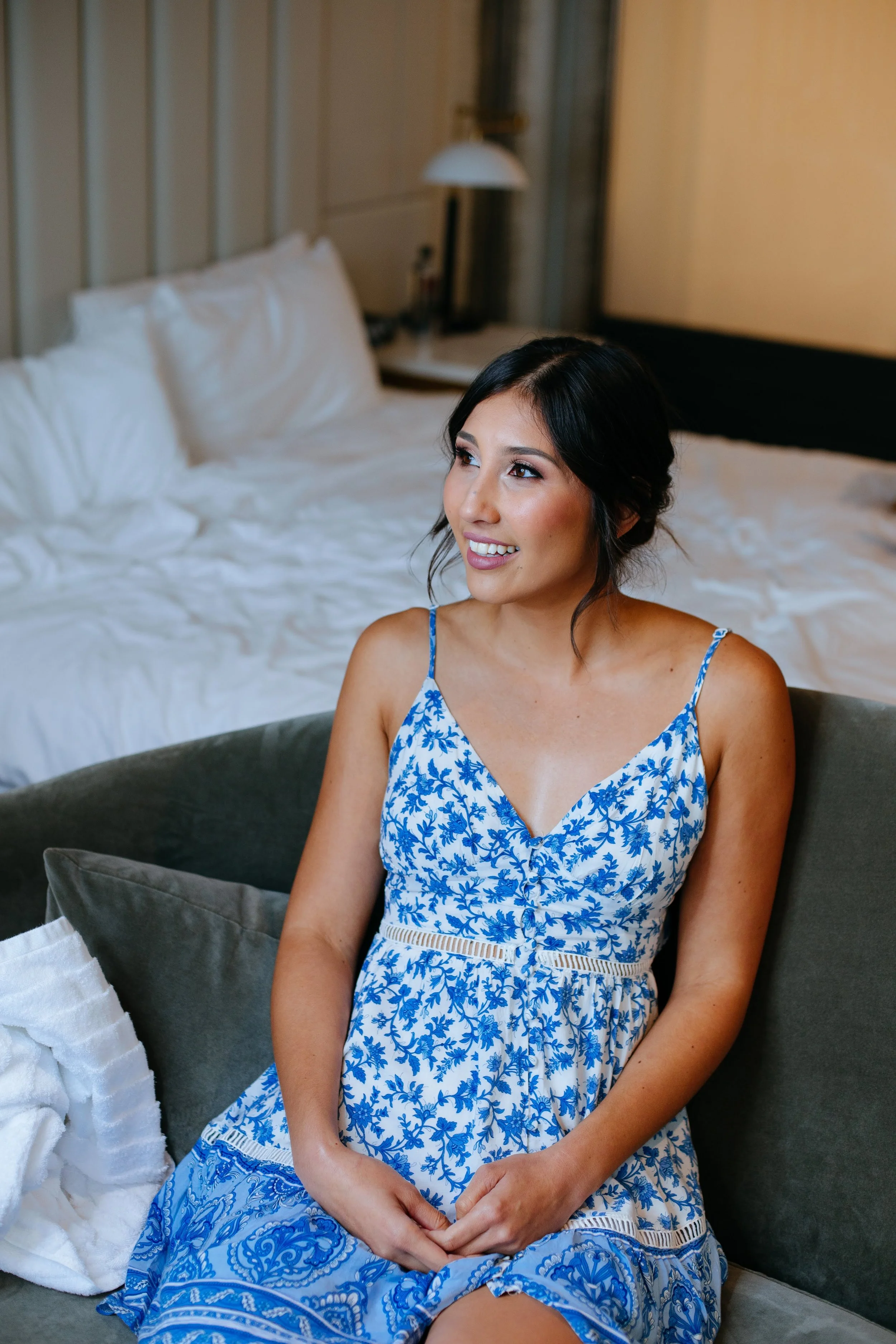 A woman with dark hair in a blue and white floral dress sitting on a green sofa in a hotel room, with a bed and bedside lamp in the background.