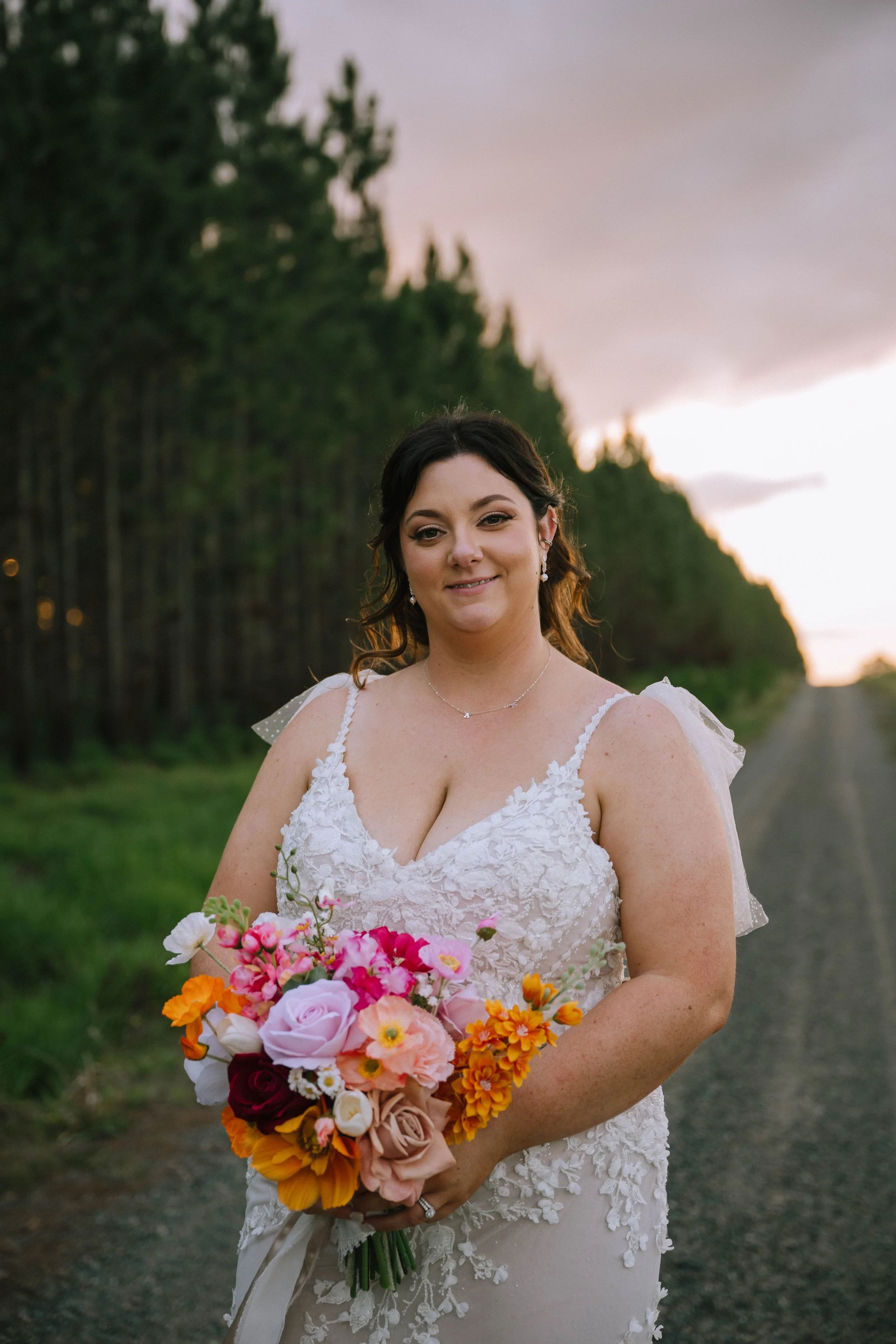A bride in a white lace wedding dress holding a bouquet of colorful flowers, standing on a rural gravel road with a forest and sunset sky in the background.