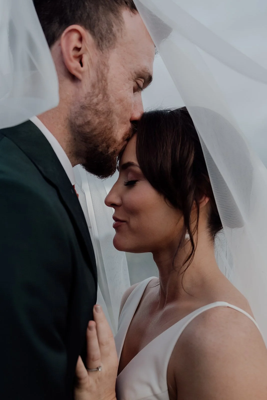 A bride and groom sharing a close, intimate moment under a white veil, with their foreheads touching and eyes closed.