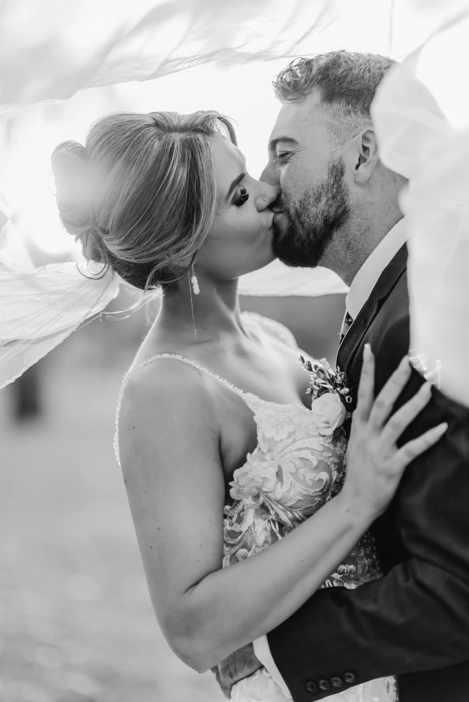 Black and white photo of a newlywed couple kissing each other, with the bride in a wedding dress and the groom in a suit.
