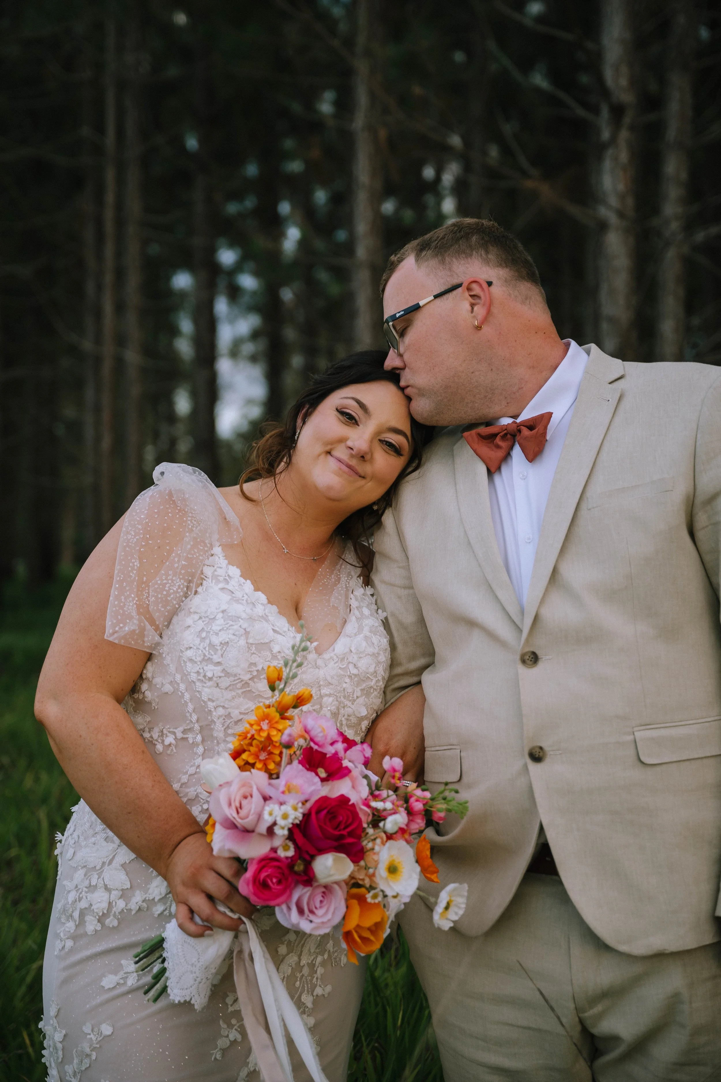 A bride and groom on their wedding day, standing close and sharing an intimate moment outdoors in a forested area. The bride holds a colorful bouquet and wears a lace wedding dress with sheer puff sleeves. The groom is dressed in a light-colored suit