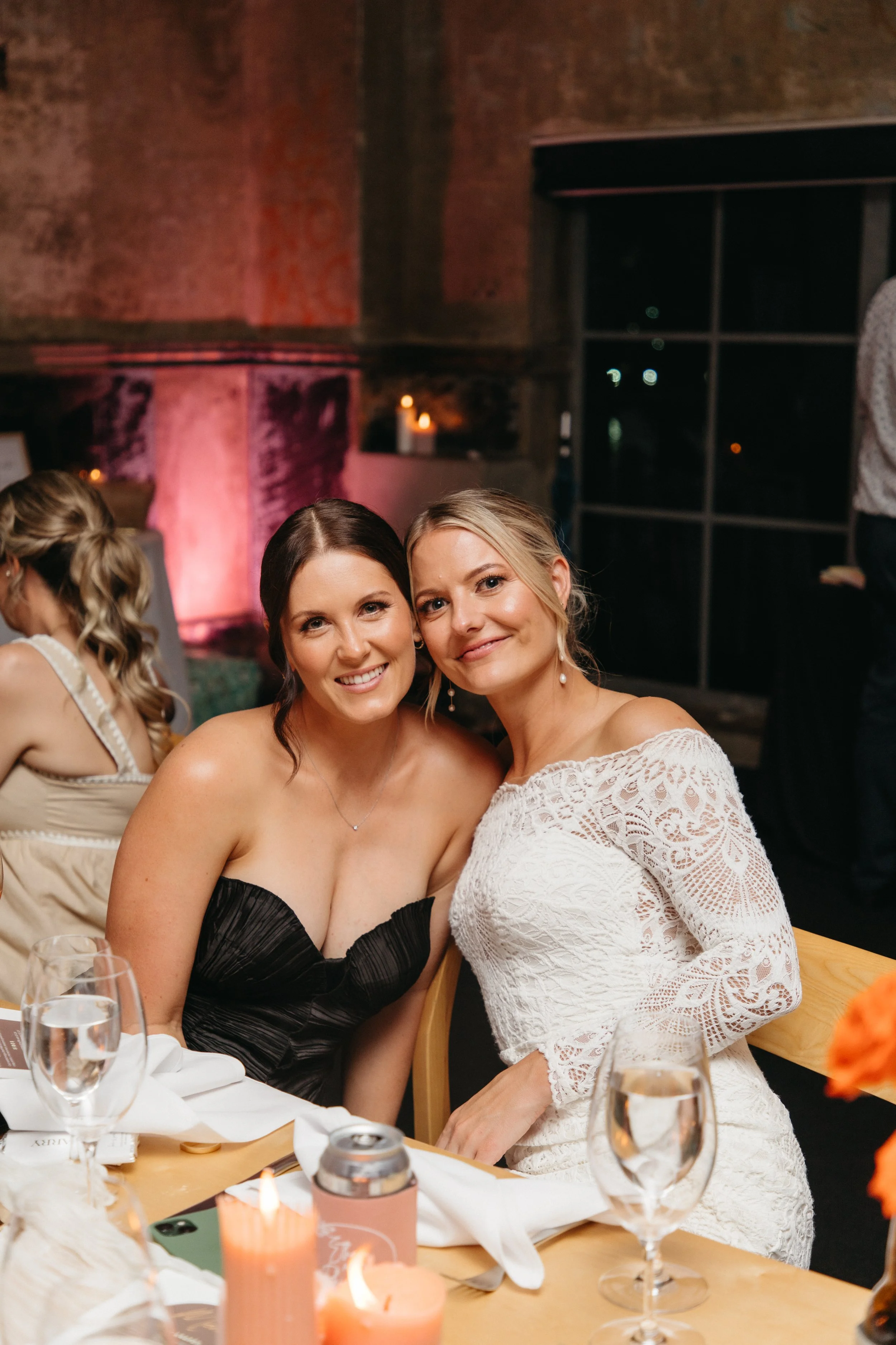 Two women sitting at a dinner table, smiling at the camera. One is wearing a strapless black dress, and the other is wearing a white lace dress with off-the-shoulder sleeves.