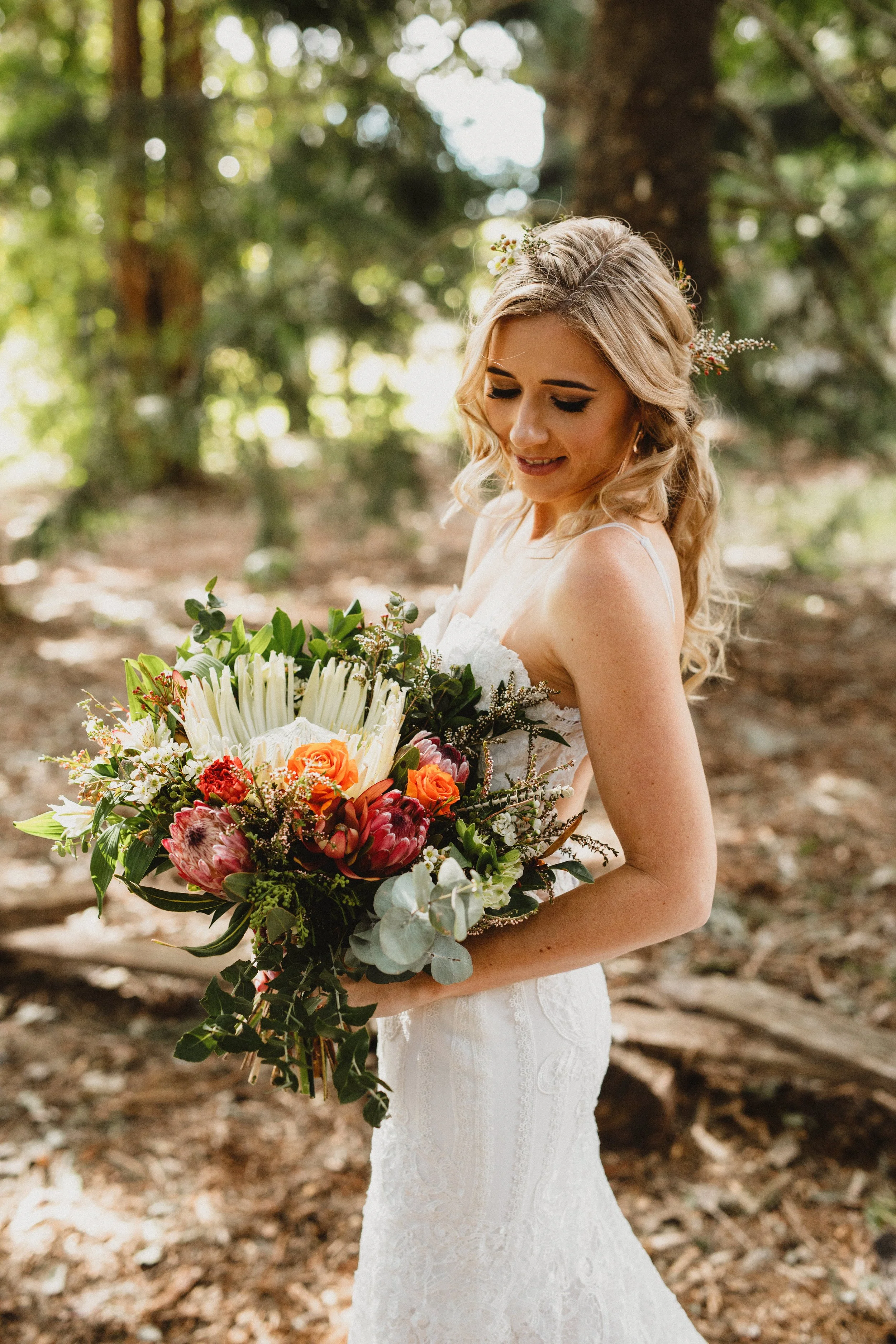 A bride in a white lace wedding dress holding a large colorful bouquet of flowers, standing in a wooded area with sunlight filtering through the trees.