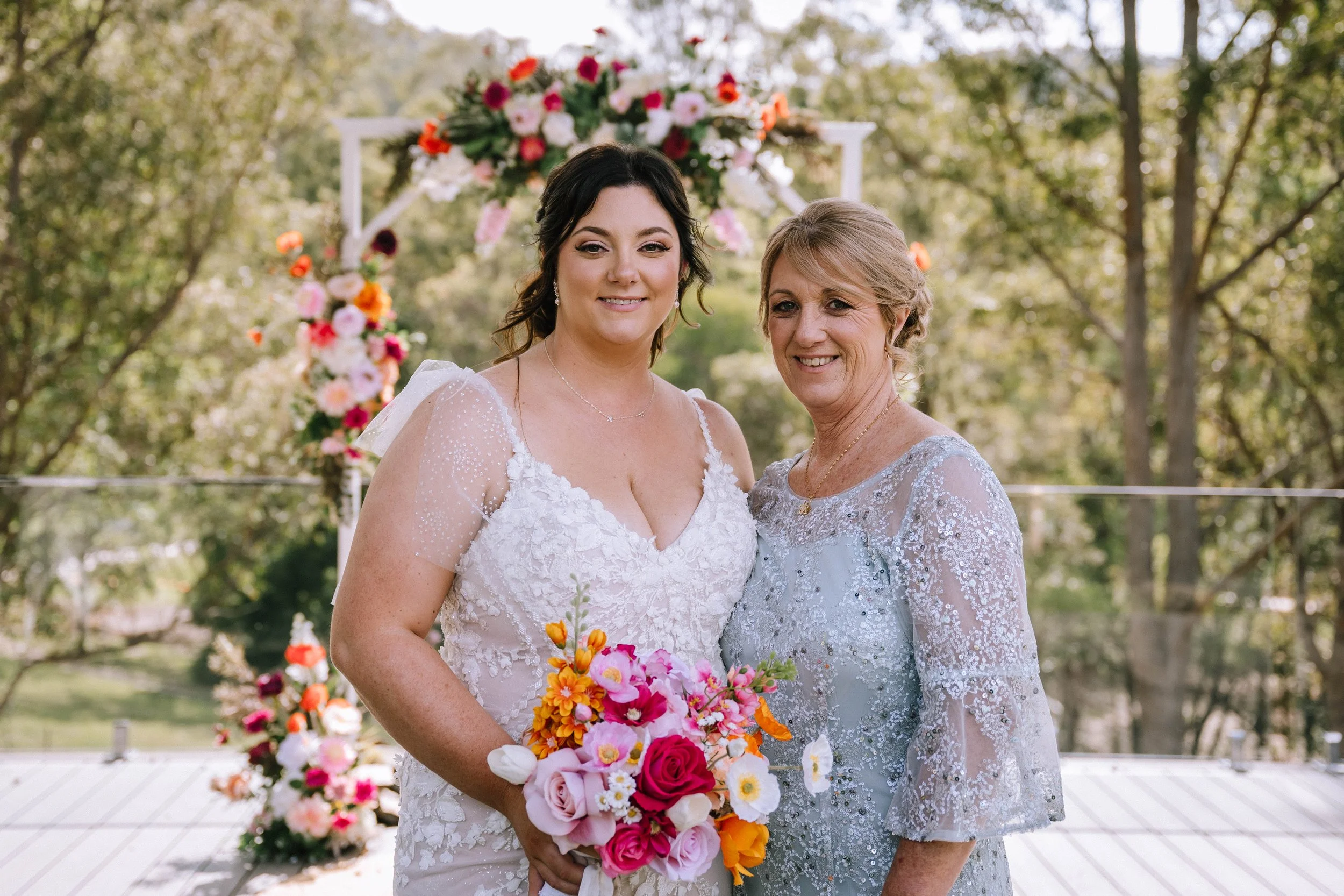 A bride in a lace wedding dress holds a vibrant bouquet of pink, orange, and white flowers, standing next to an older woman in a light blue lace dress, at an outdoor wedding venue with floral archway in the background.