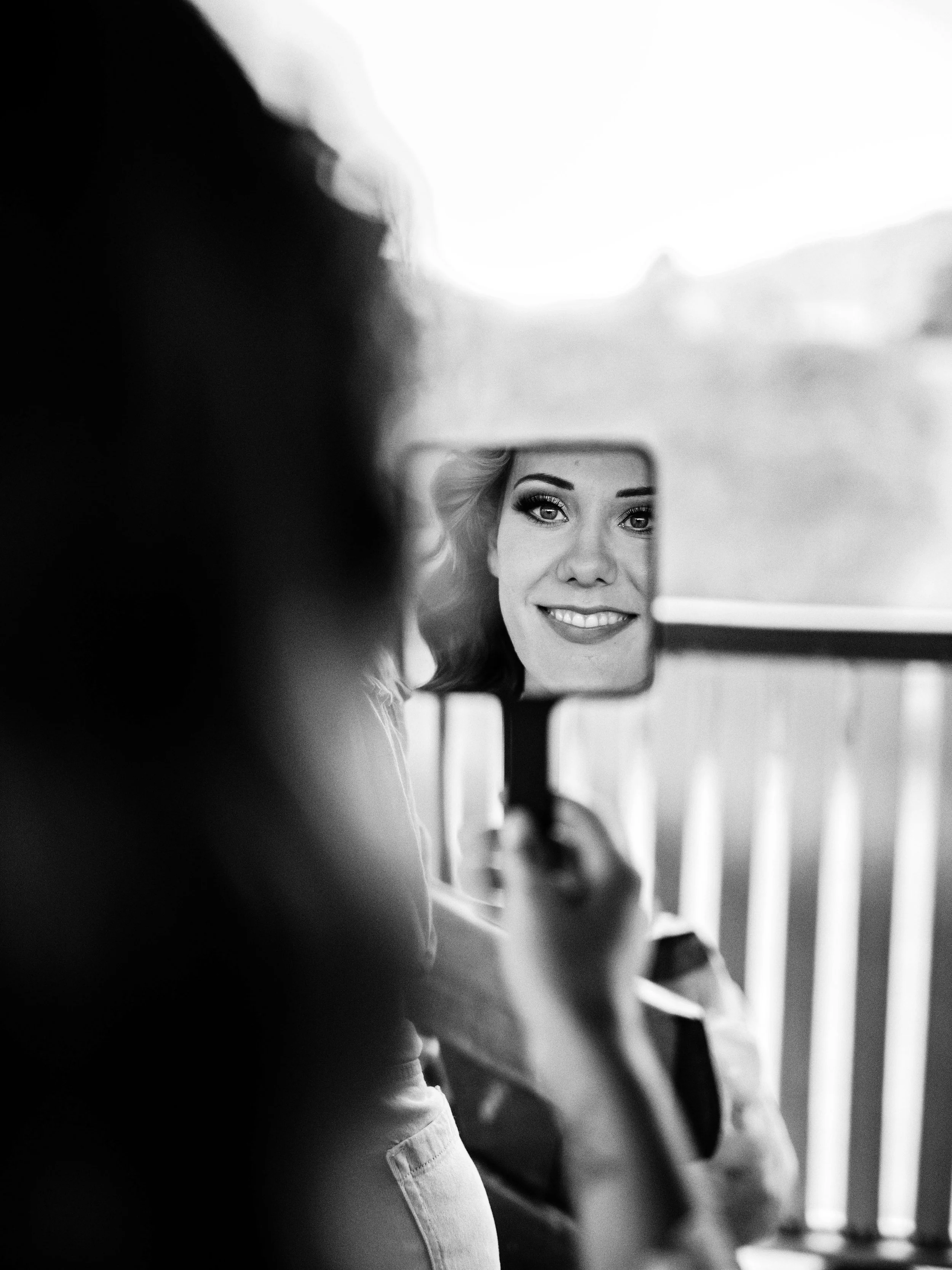 A woman smiling at her reflection in a mirror while holding it up to her face, black and white photograph.