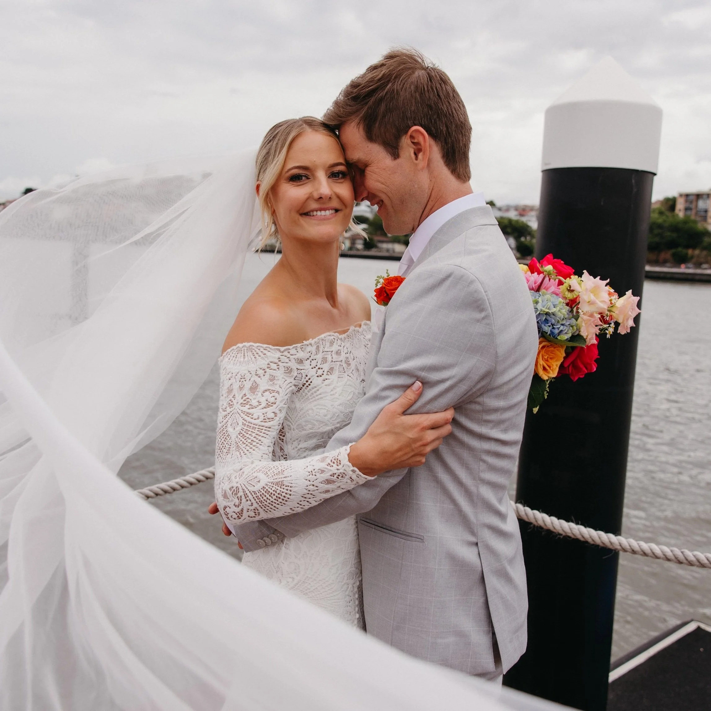 A newlywed couple on a dock near a body of water, embracing and smiling, with the bride wearing a lace off-shoulder wedding dress and veil, and the groom in a light gray suit holding a colorful bouquet of flowers.