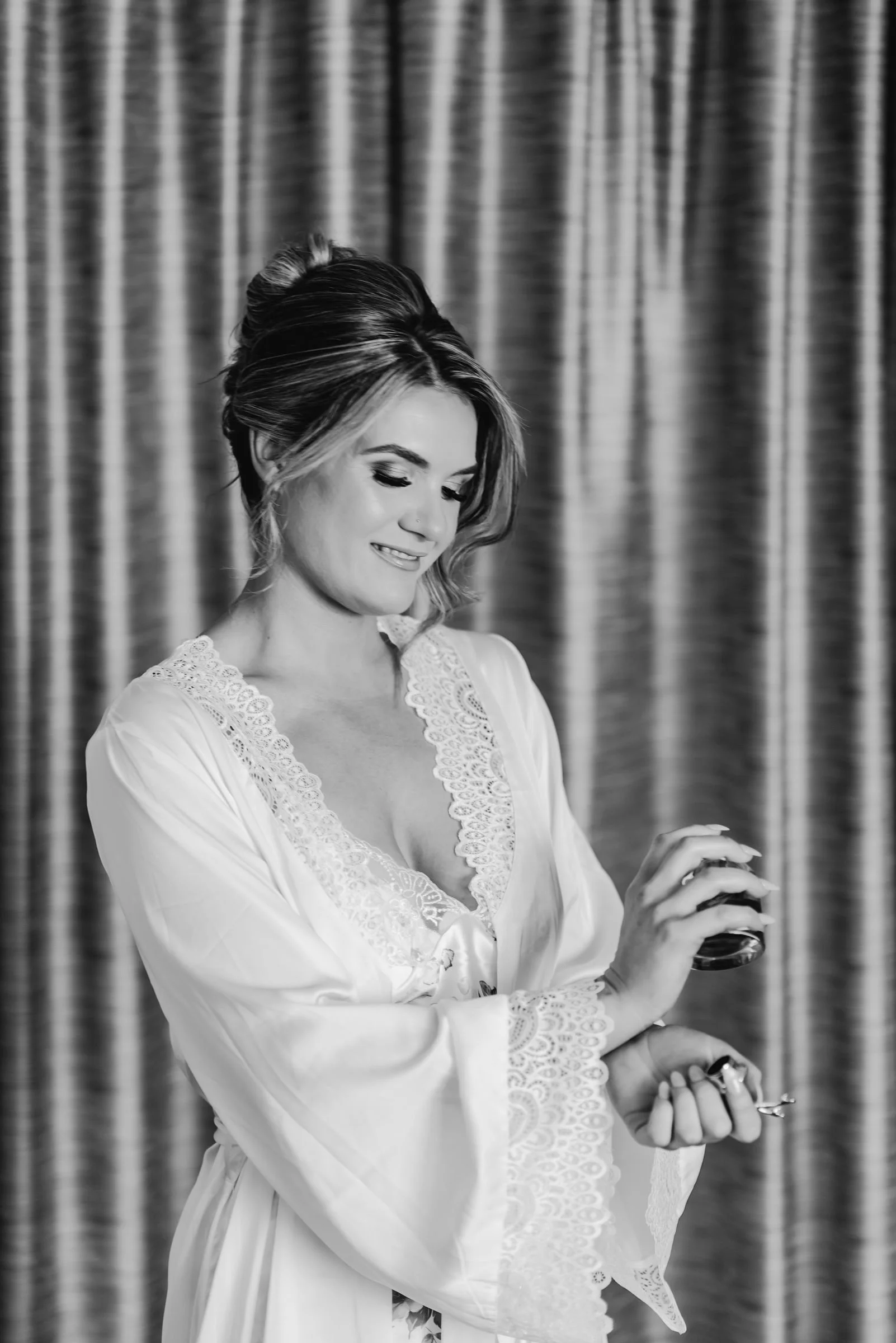 A woman in a satin robe with lace details, adjusting her ring while holding a small perfume bottle, smiling with her eyes down, in front of a curtain.