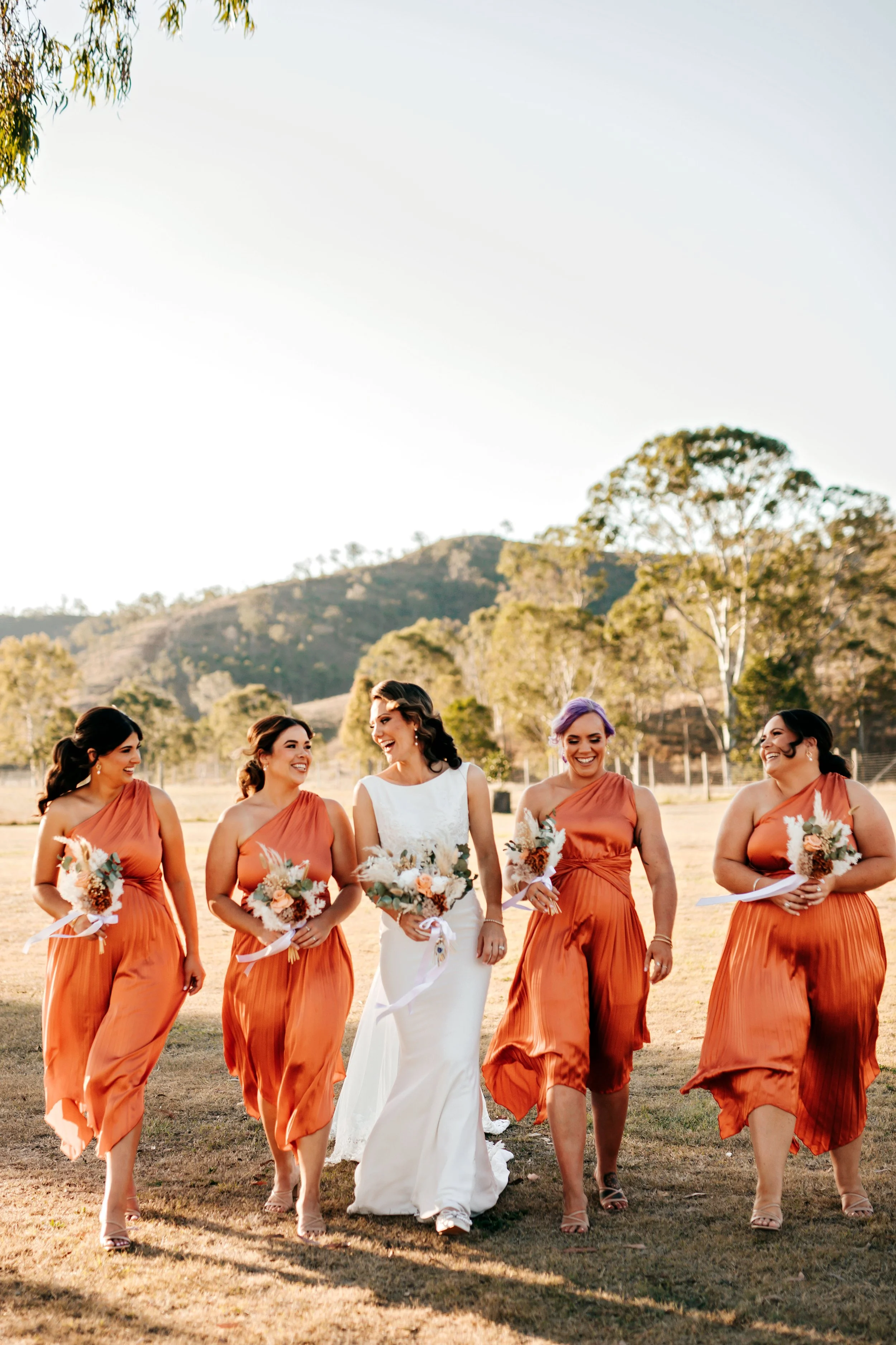 A bride and four bridesmaids walking outdoors on a sunny day, holding bouquets, with trees and hills in the background.