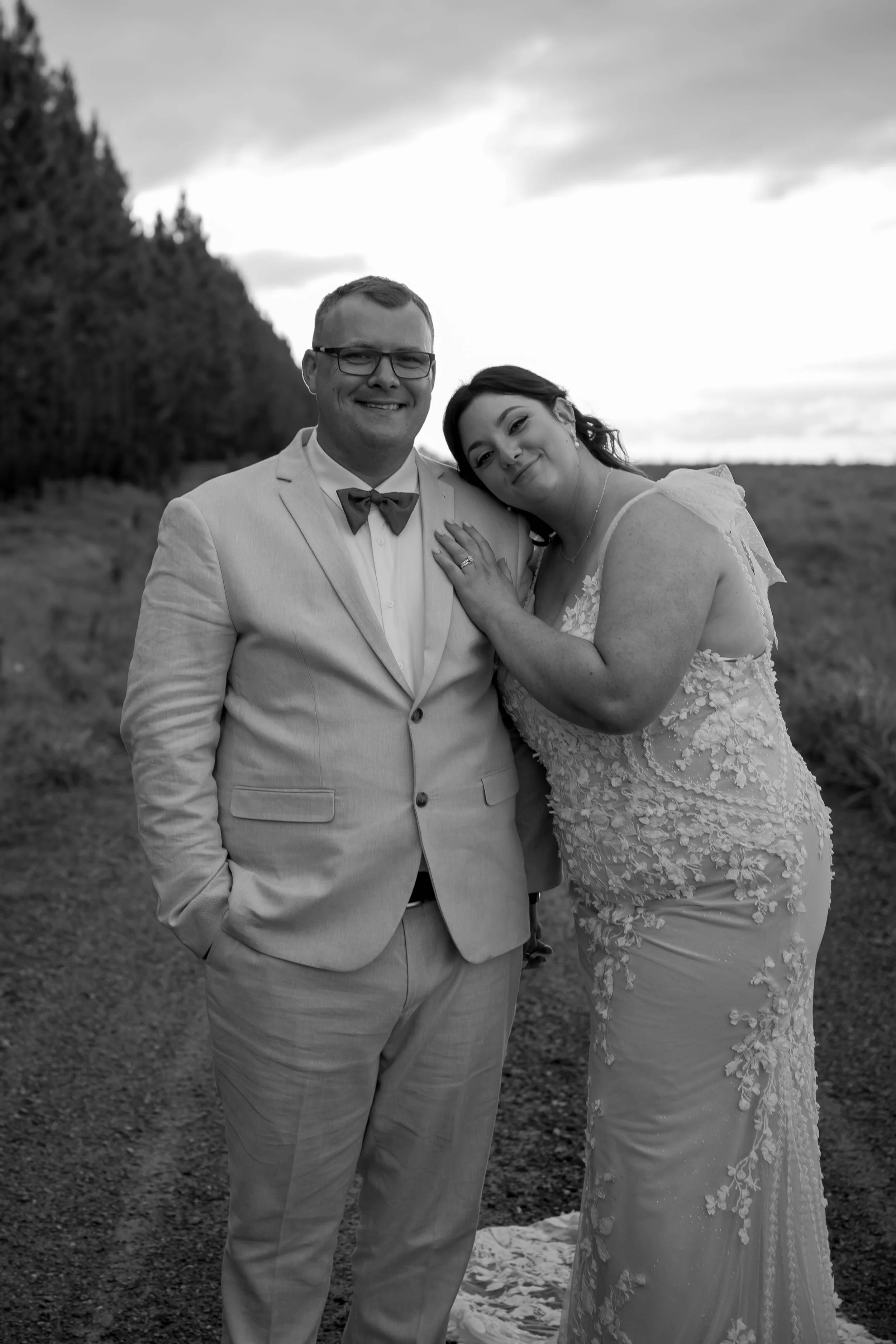 Black and white photo of a bride and groom standing outdoors, smiling, with the bride resting her hand on the groom's chest, in a natural setting with trees and open sky in the background.