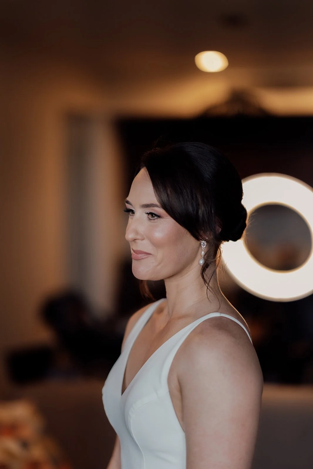 A woman dressed in a white dress with dark hair styled in an elegant updo, smiling softly, with a ring light behind her creating a circular glow in the background.