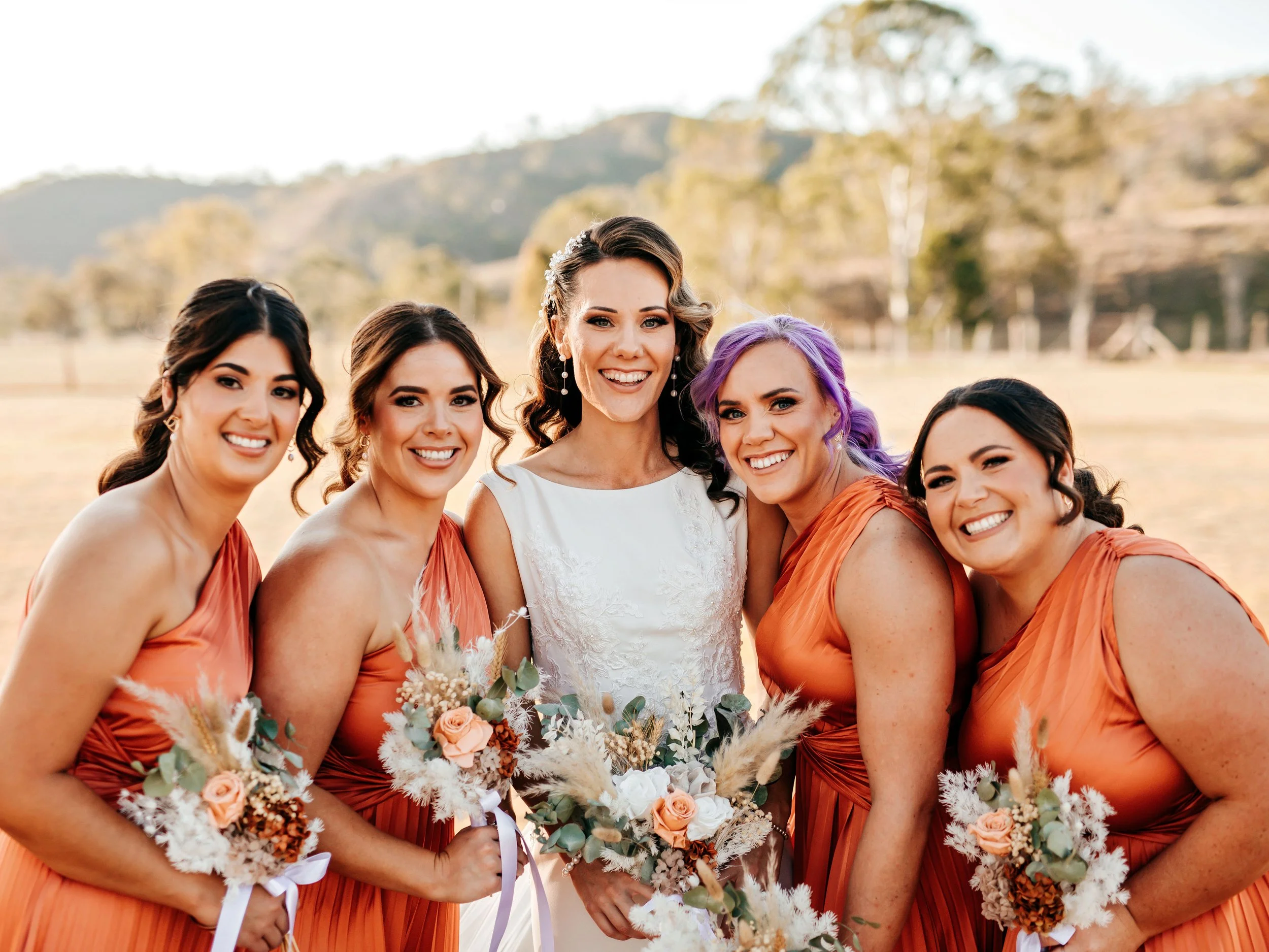 A bride in a white wedding dress with brunette hair standing with five bridesmaids dressed in orange dresses, outdoors on a sunny day with trees and hills in the background. They are smiling and holding bouquets of flowers.