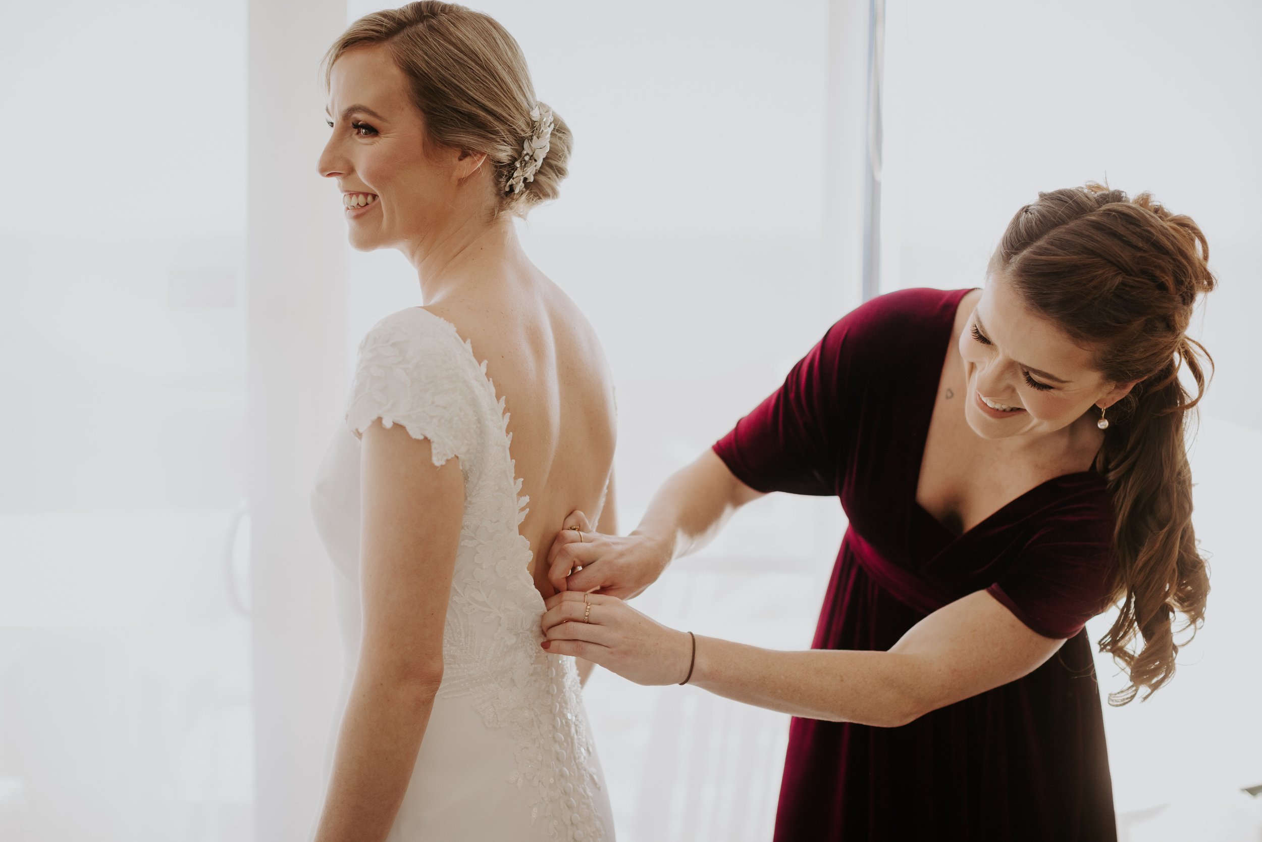 A bride in a white wedding dress is getting help from a woman in a burgundy dress to button up her dress from behind.