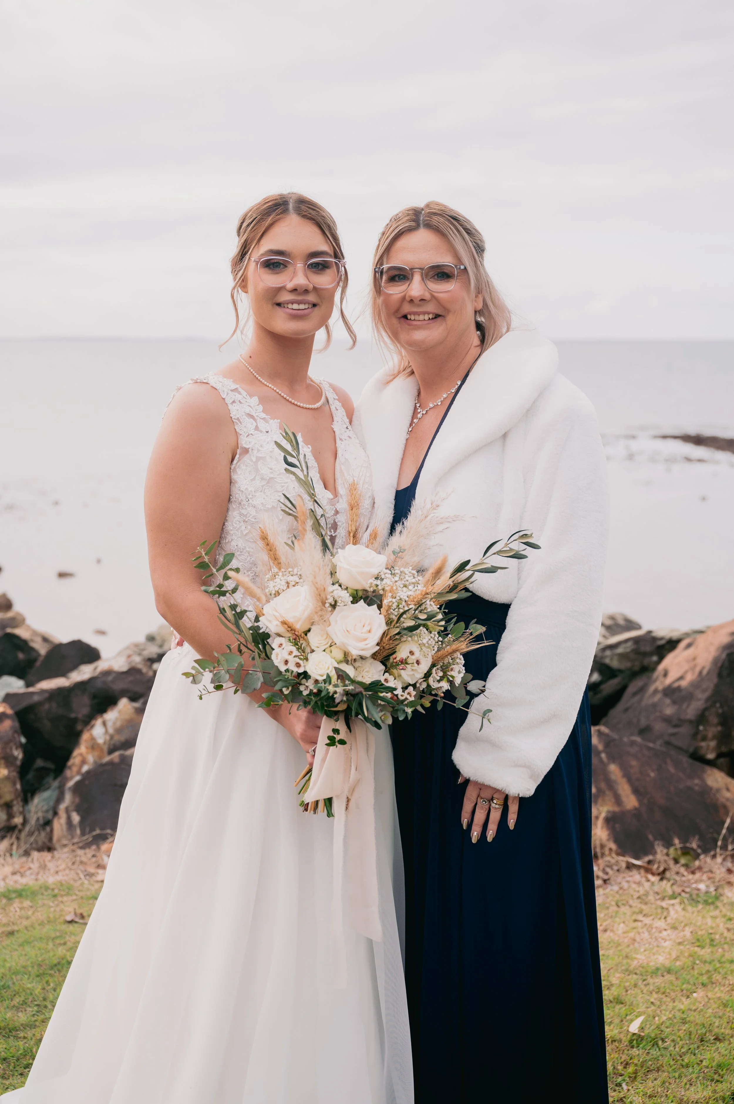 A bride and an older woman, possibly her mother, standing outdoors near the water on a cloudy day, smiling and holding a bouquet of white roses and greenery.