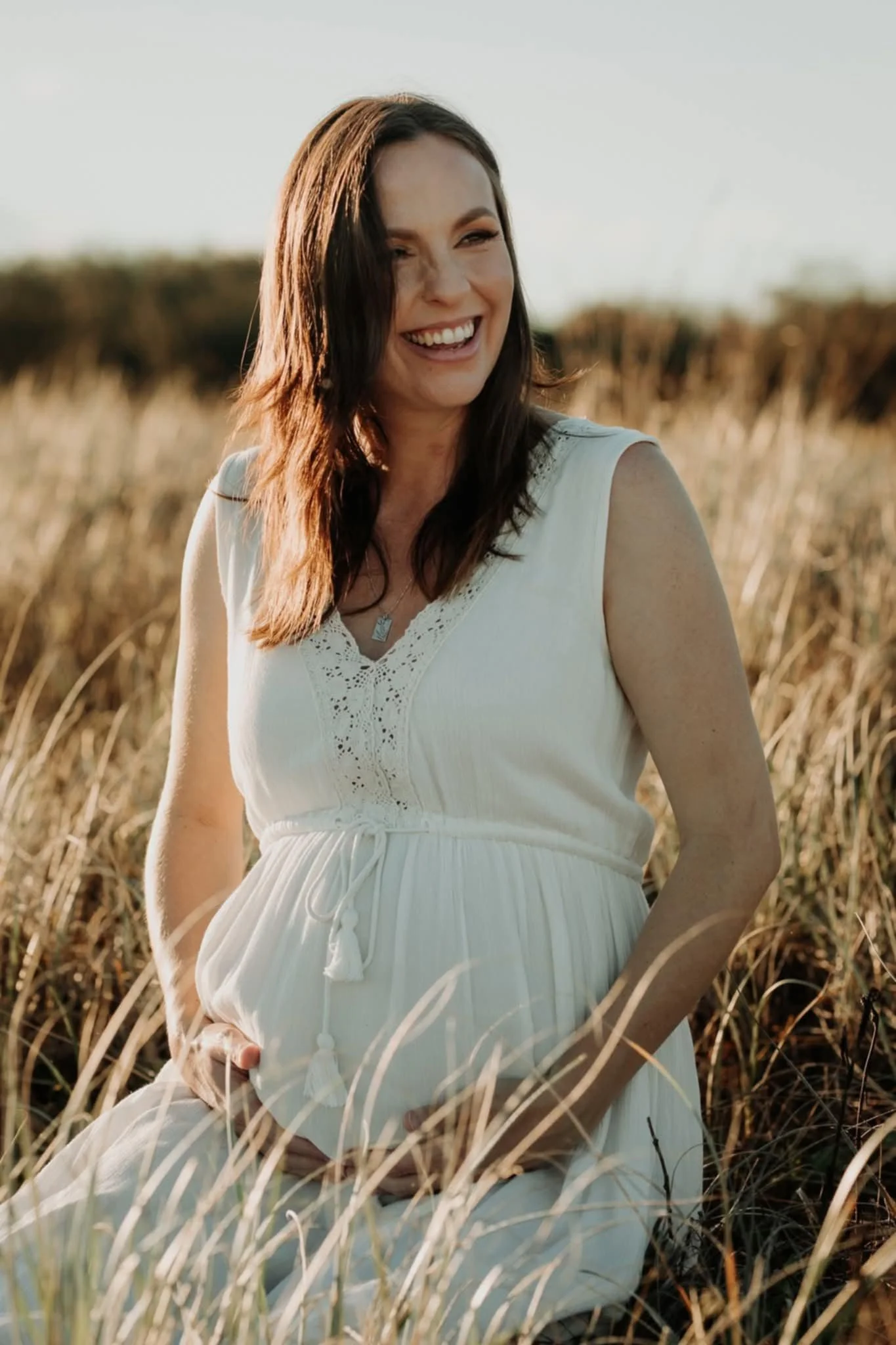 A woman with brown hair smiling and holding her pregnant belly while kneeling in a field of tall grass during sunset.