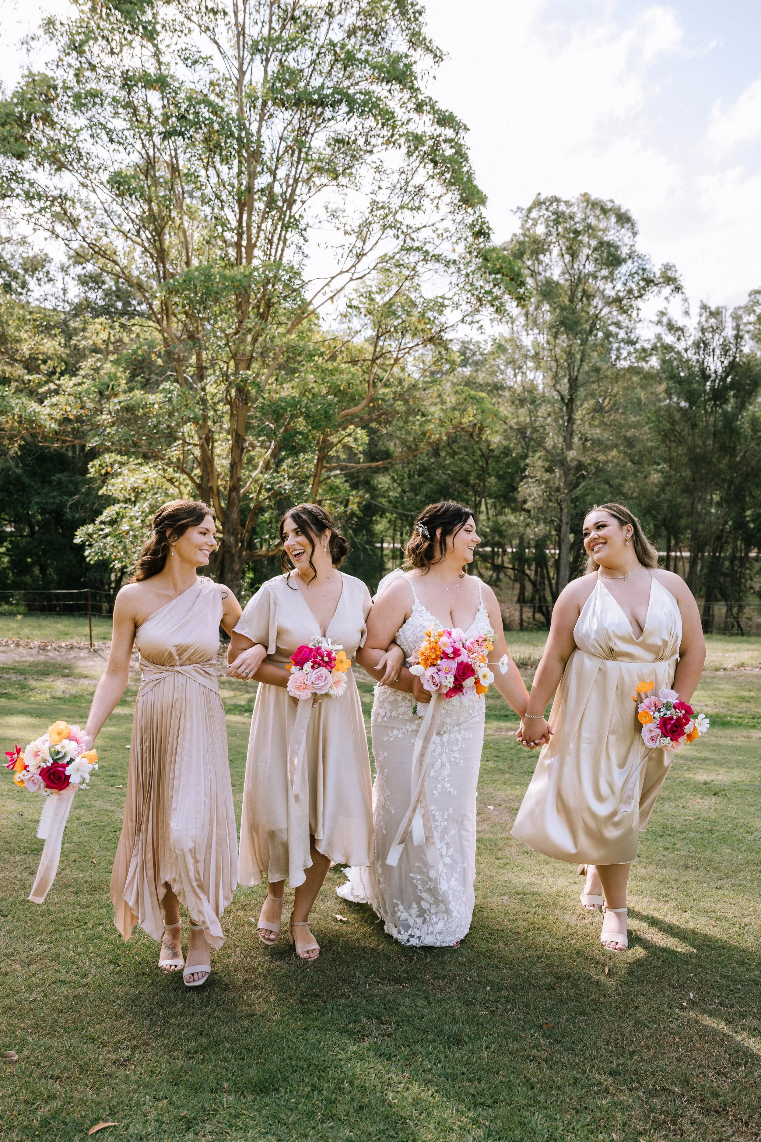 A group of four women, dressed in elegant attire, walking on a grassy field holding colorful bouquets, with trees and a cloudy sky in the background.