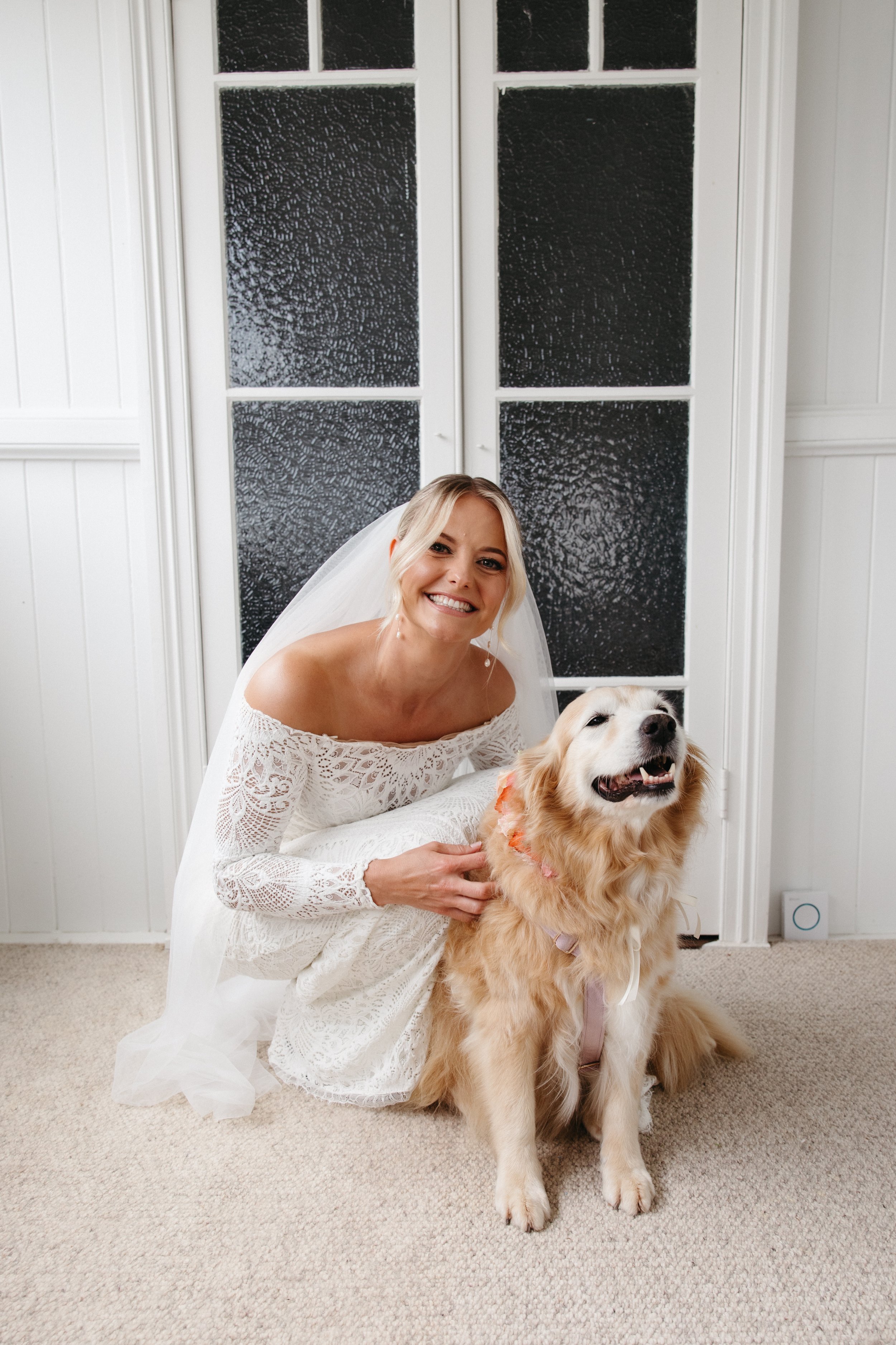 A smiling bride in a white lace wedding dress kneeling next to a golden retriever, both inside a room with white paneled walls and a glass-paneled door in the background.