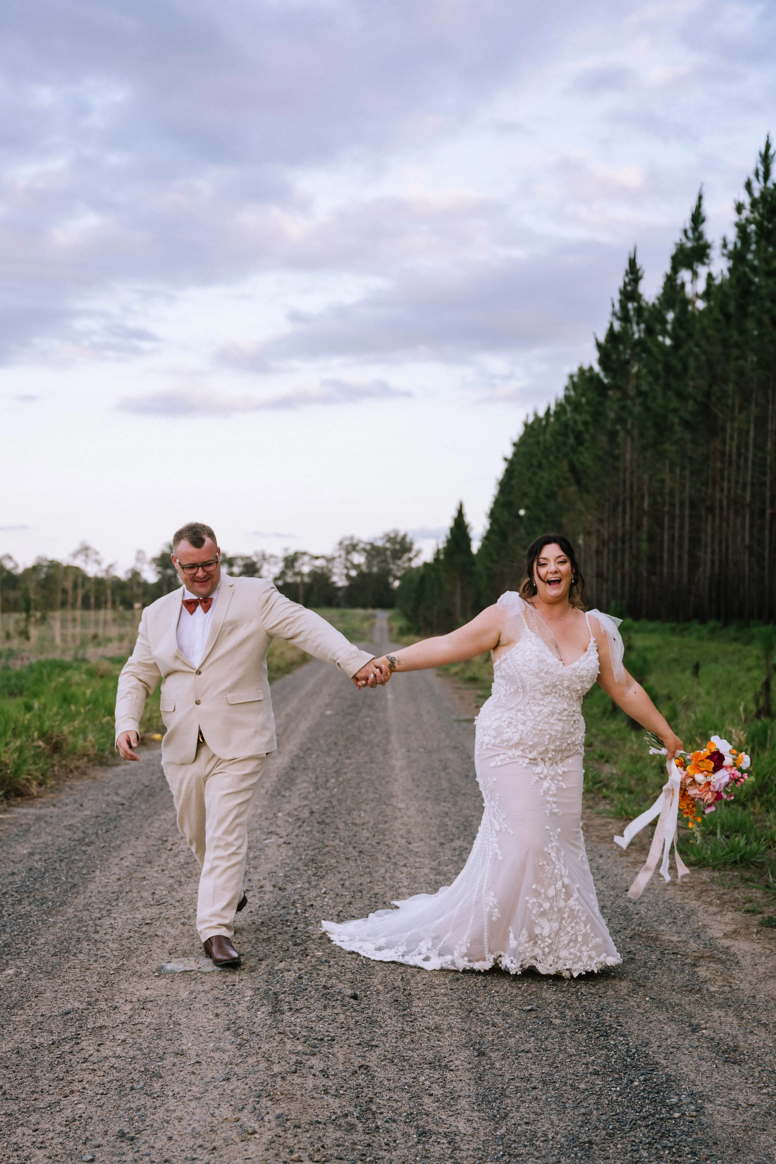 Happy couple in wedding attire walking hand in hand on a gravel road in a rural area, with trees and a cloudy sky in the background.