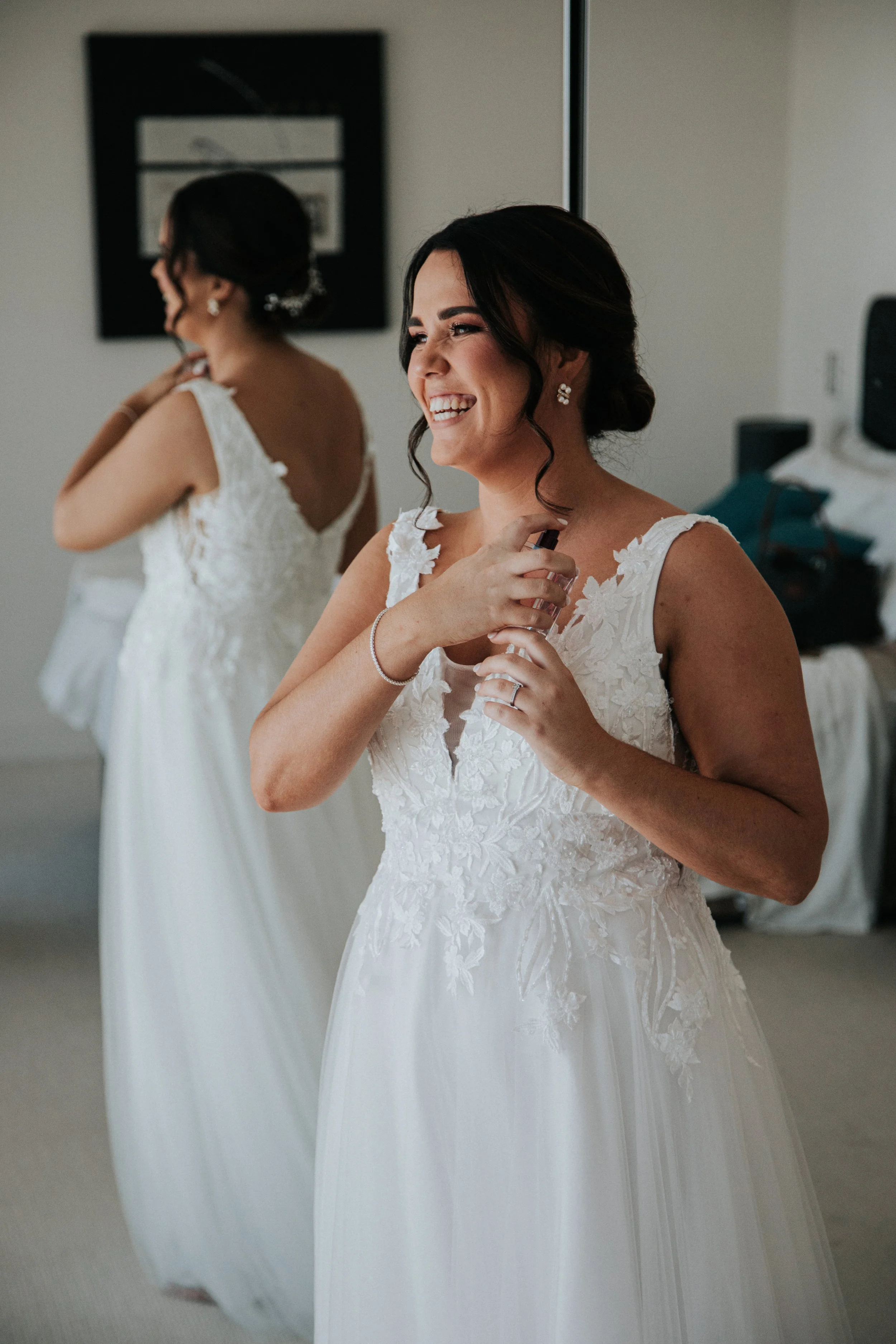 A smiling bride in a white lace wedding dress holding a perfume spray in a dressing room, with her reflection visible in a mirror behind her.