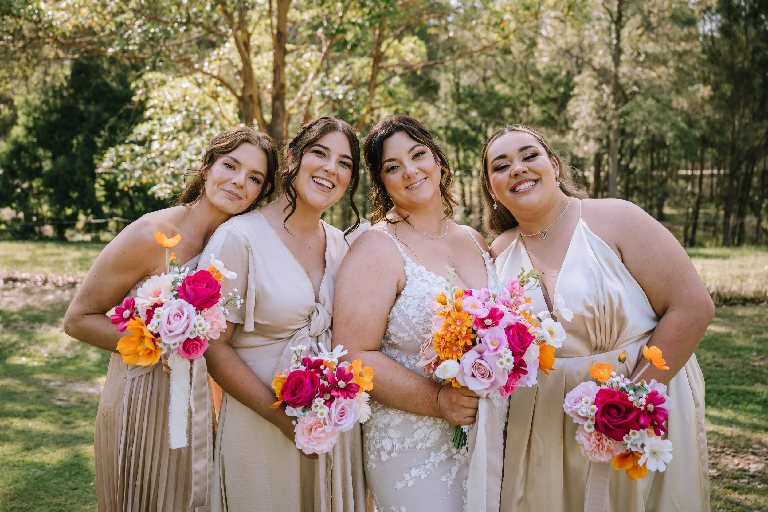 A group of four women at a wedding, standing outdoors in a park-like setting with greenery and trees in the background. The bride, in a white lace wedding dress, is holding a colorful bouquet, and three bridesmaids in light tan dresses are holding ma