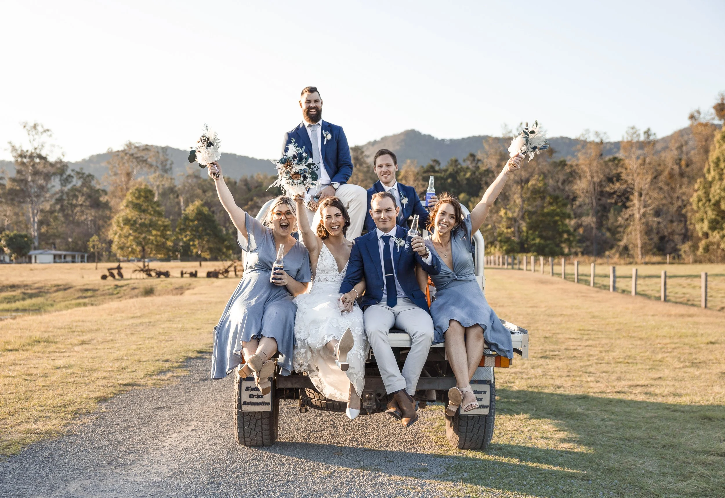 Happy wedding party sitting on a truck outdoors with mountains in the background. The bride and groom are seated in the center, smiling, with bridesmaids and groomsmen around them, some standing and others sitting, celebrating with drinks and flowers