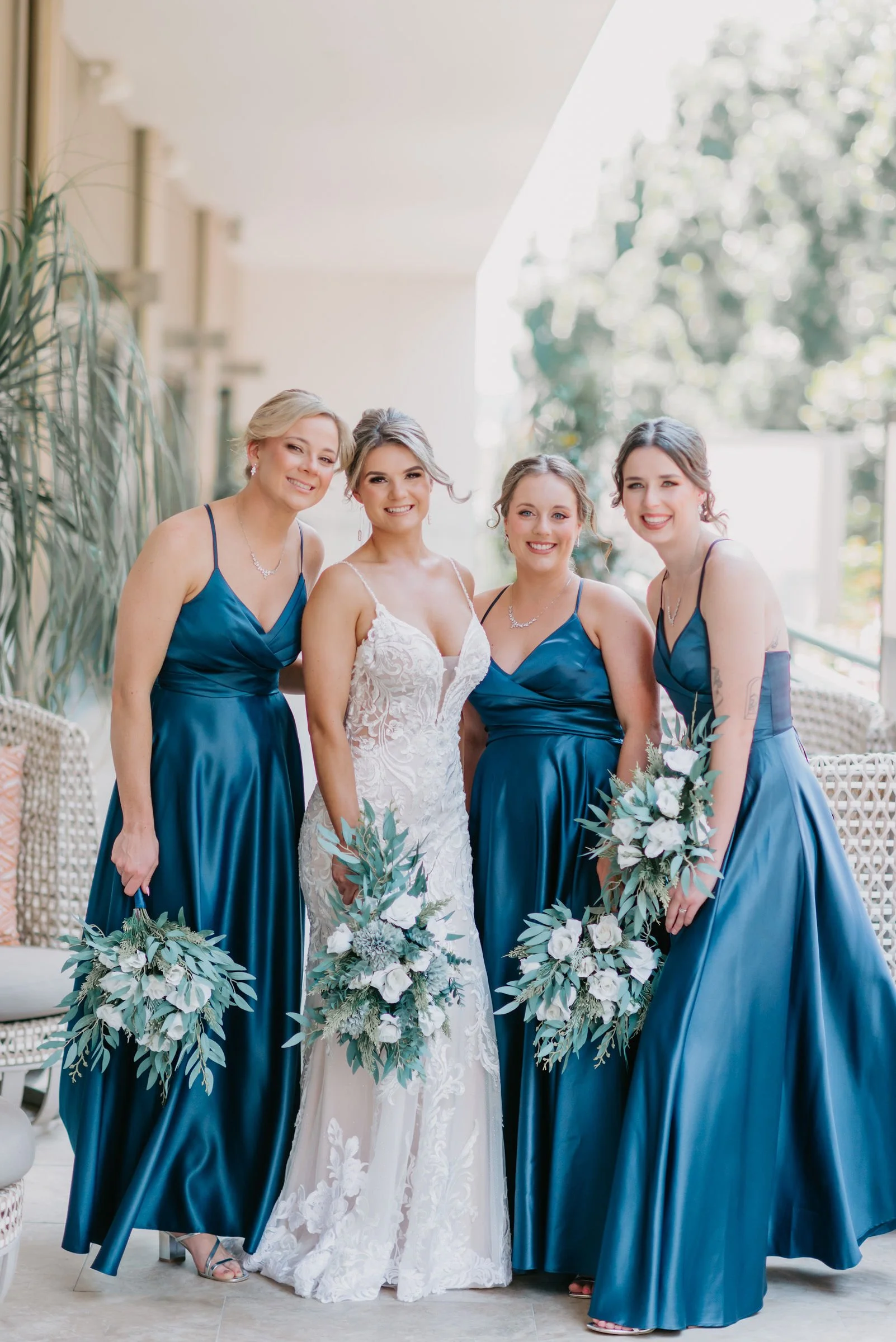Four women in formal dresses, one in a white wedding gown and three in matching blue bridesmaid dresses, standing together, smiling, holding bouquets, on a light-colored porch with greenery in the background.