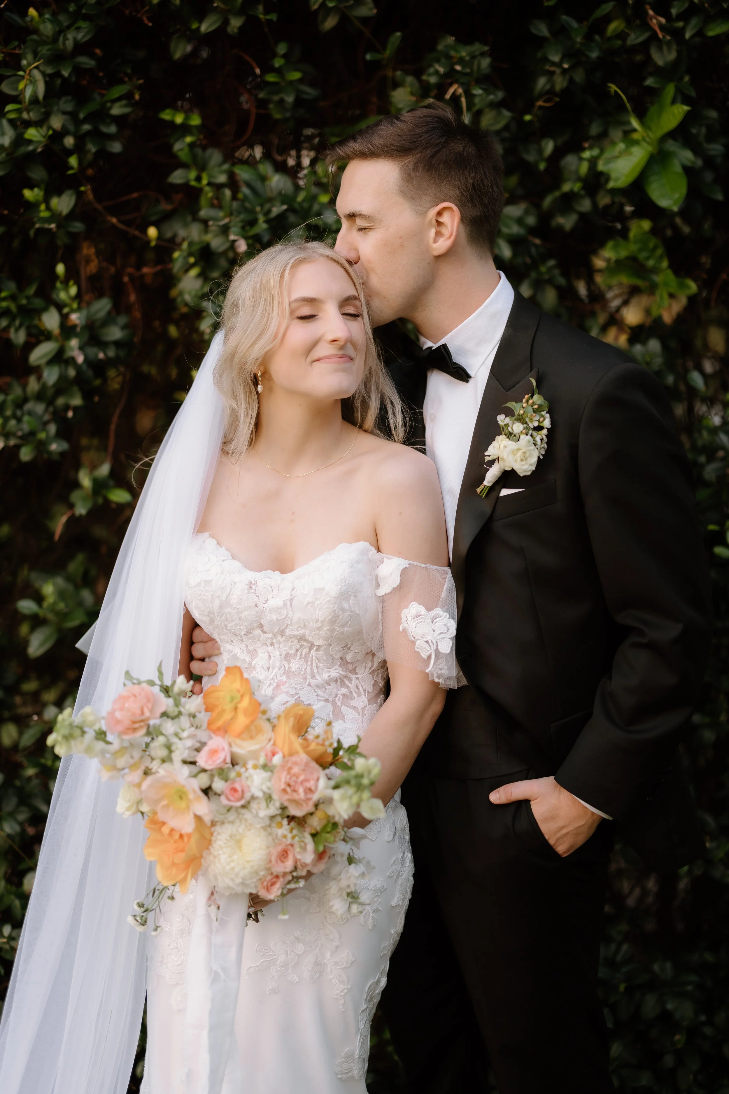 A bride and groom share a tender moment outdoors, with the groom kissing the bride's forehead. The bride has blonde hair, a white lace wedding dress, and holds a bouquet of pink, white, and peach flowers. The groom is dressed in a black tuxedo with a