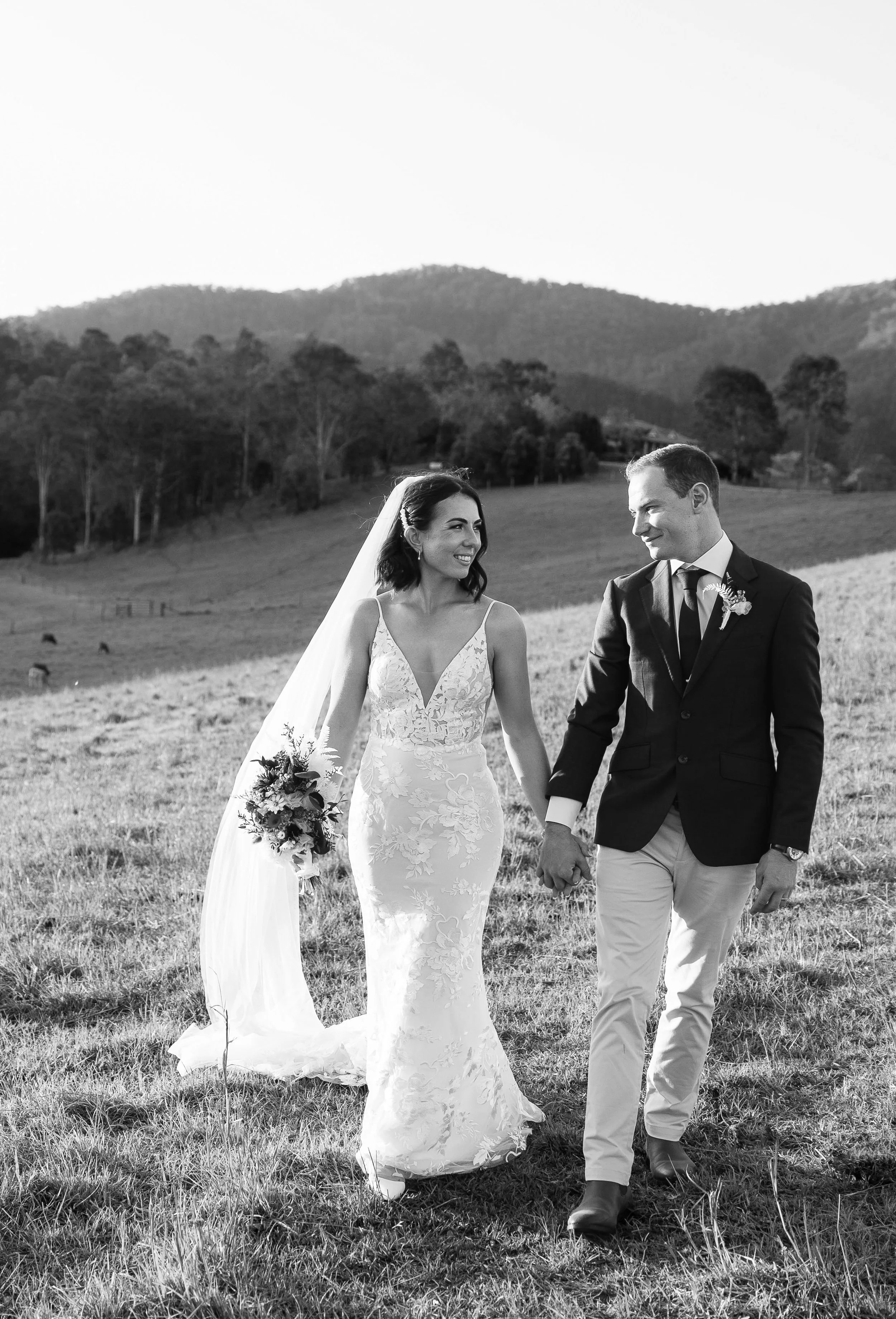 Black and white photo of a bride and groom holding hands, walking in a grassy field with trees and mountains in the background, wedding attire and smiling at each other.