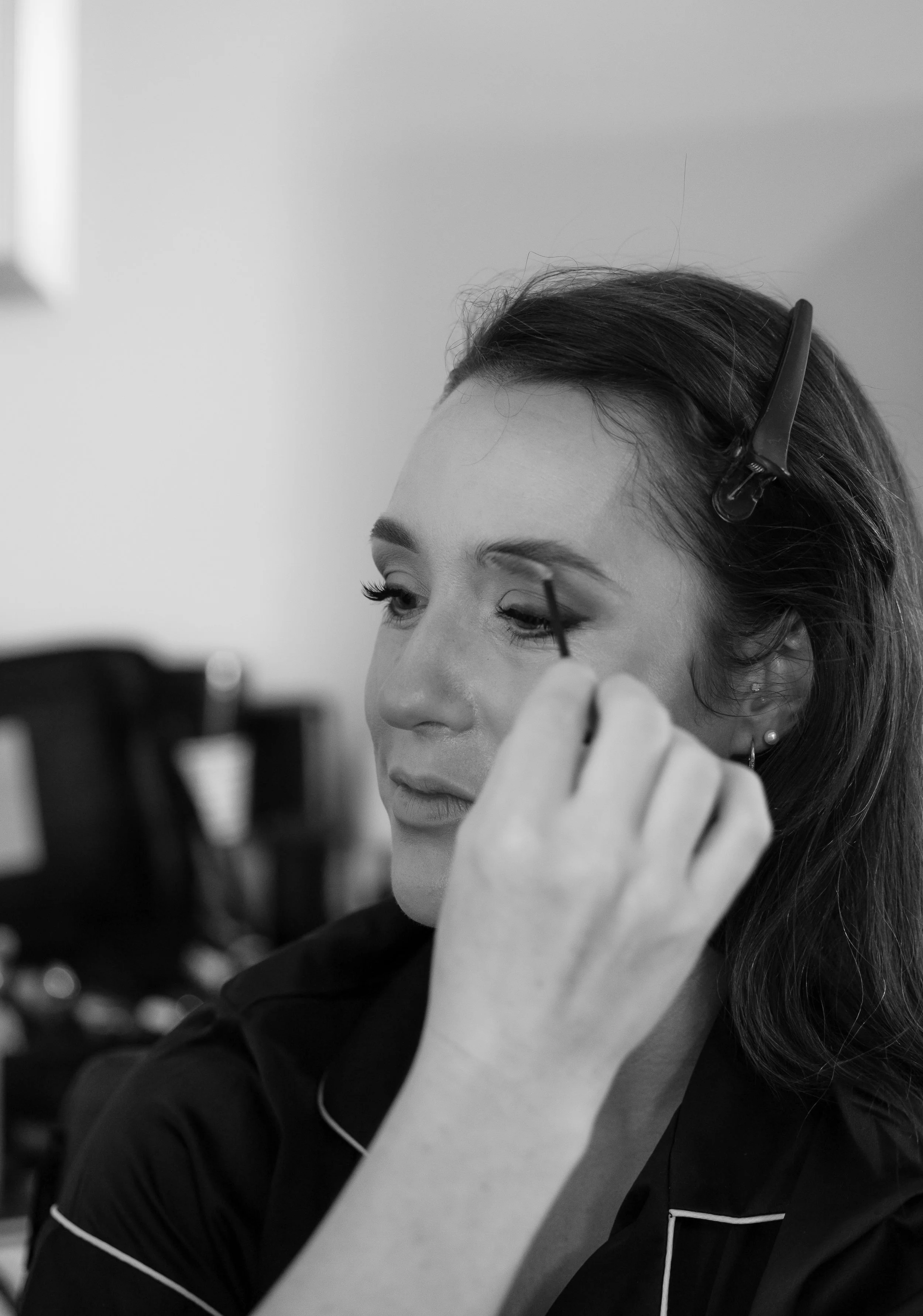 A woman with a hair clip applying makeup, specifically eyeliner, in front of a mirror.