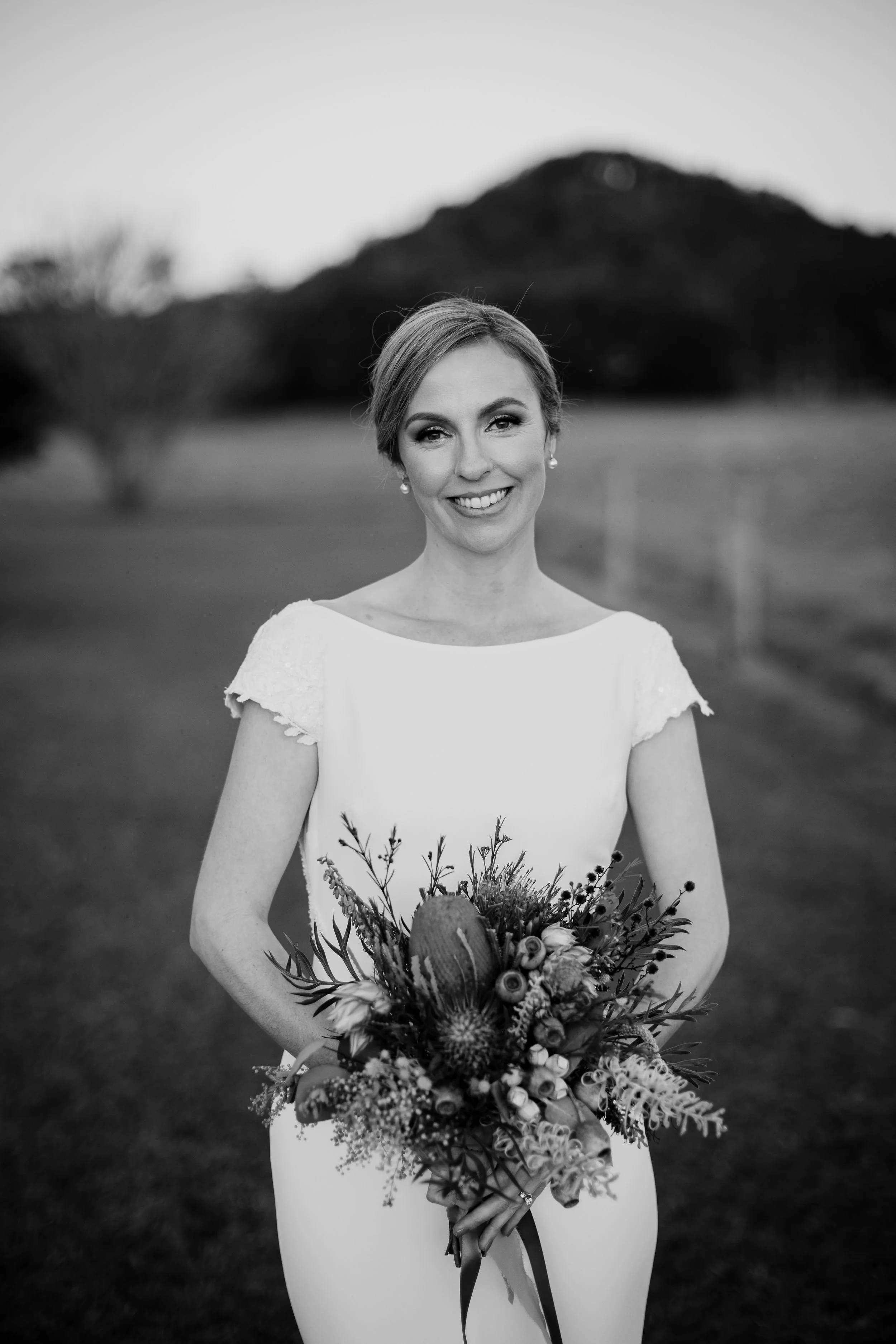 Black and white photo of a woman in a wedding dress, holding a bouquet of flowers, standing outdoors with trees and a hill in the background.