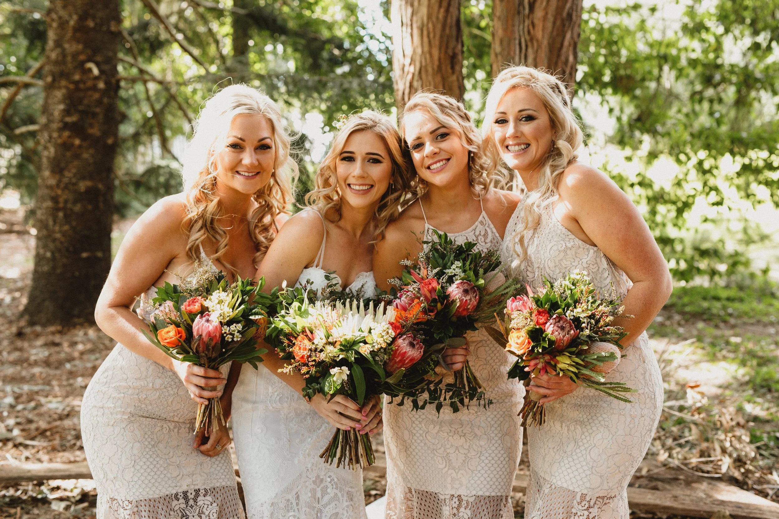 Four smiling women in white lace dresses holding bouquets, standing outdoors among trees