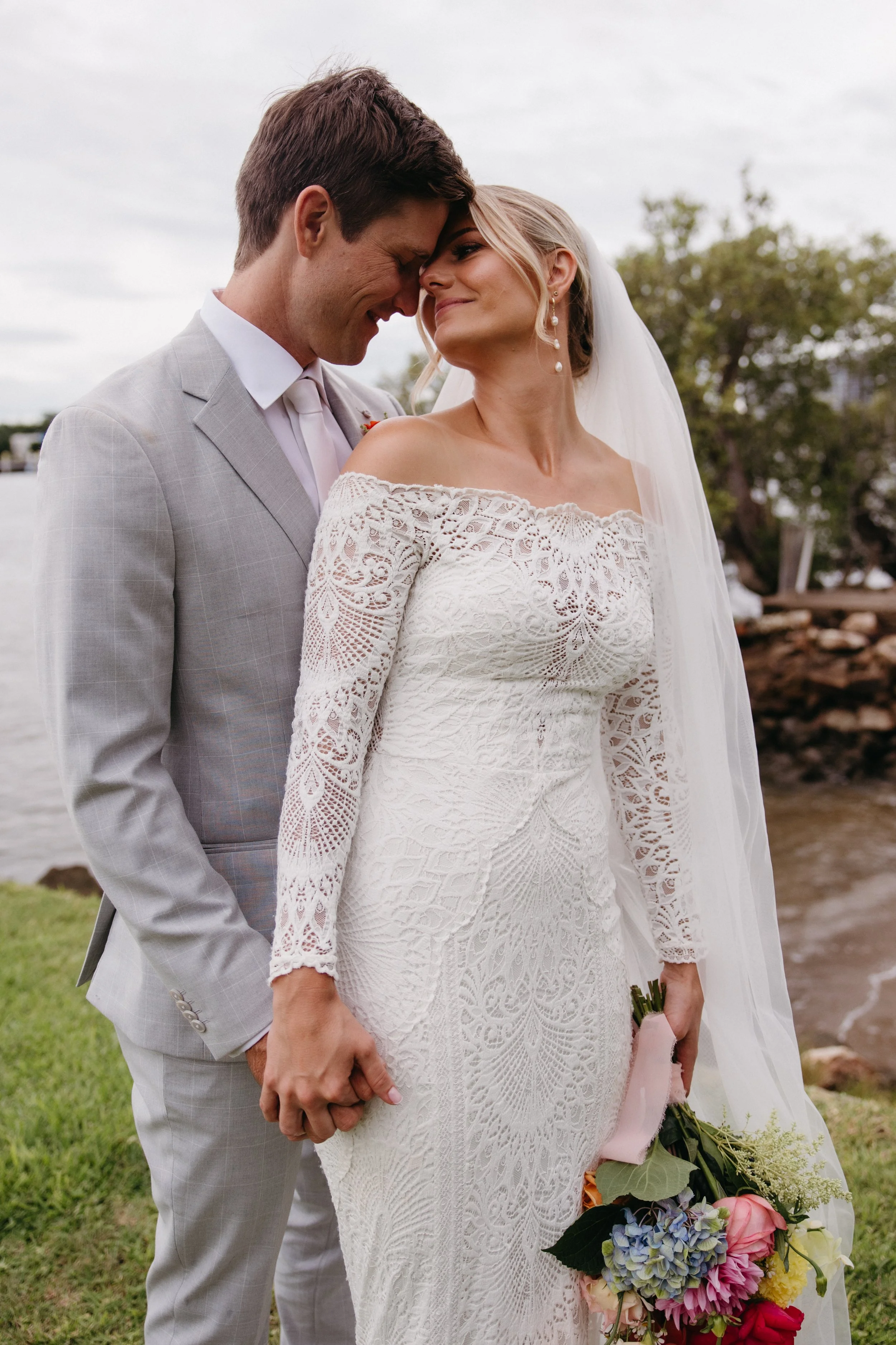 A bride and groom sharing an intimate moment outdoors near a body of water, with the groom in a light gray suit and the bride in a lace off-shoulder wedding dress holding a colorful bouquet.