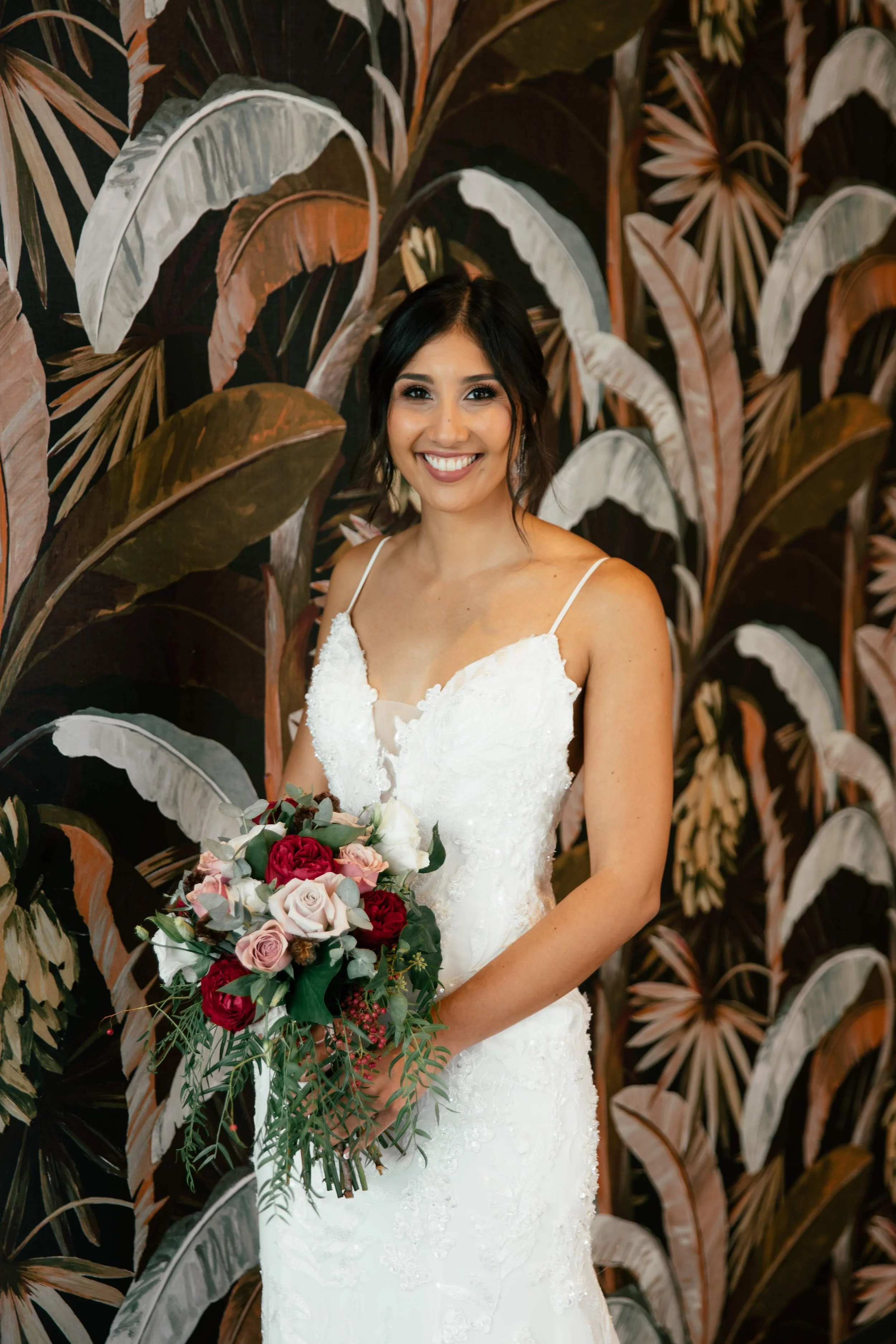 A smiling bride in a white wedding gown holding a bouquet of pink, red, and white roses, standing in front of a tropical leaf-patterned wall.