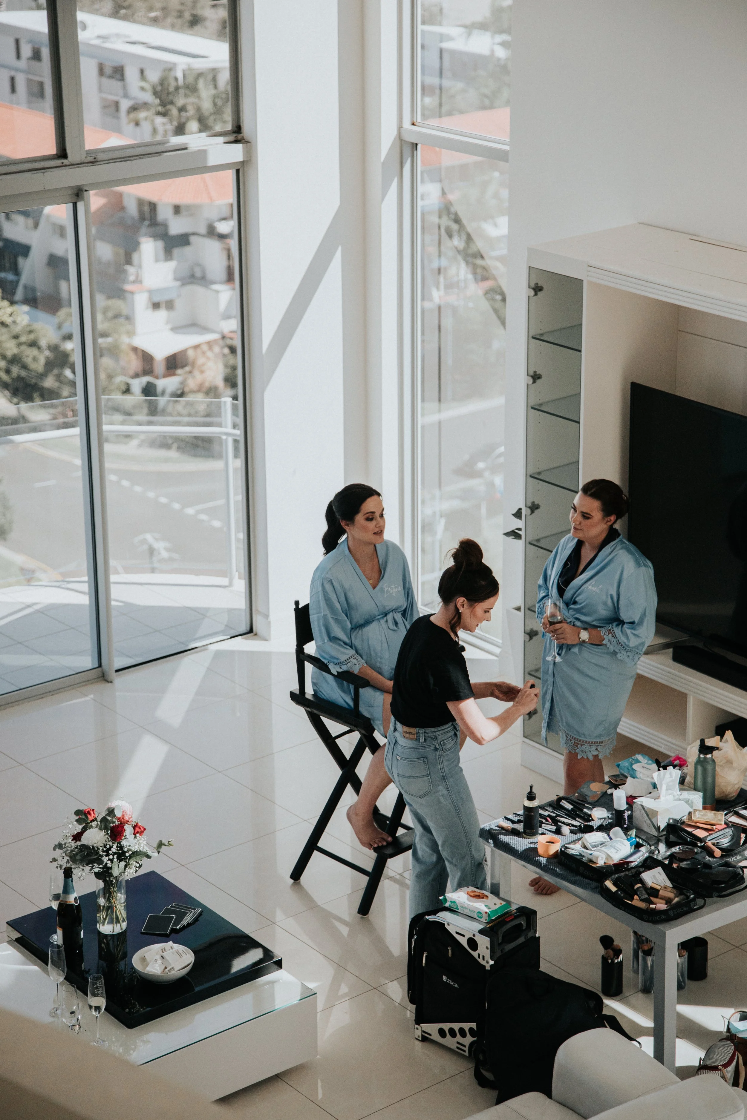 Three women in blue bathrobes and a woman in a black shirt and jeans preparing makeup and hair at a bright, modern living room with large windows, a TV, a table with makeup supplies, and flowers.