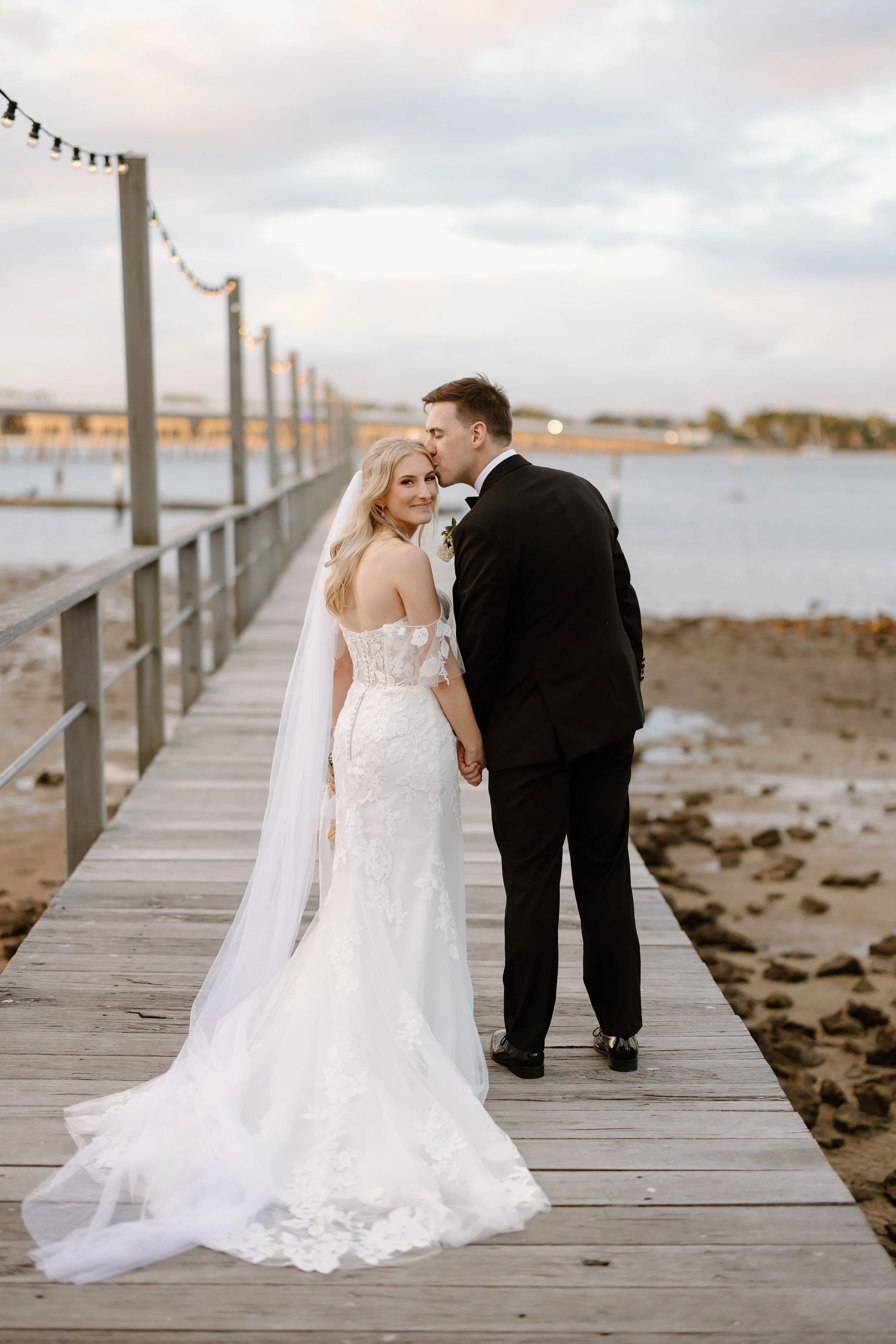 A bride and groom holding hands on a pier by the water during sunset, with the groom kissing the bride's forehead.