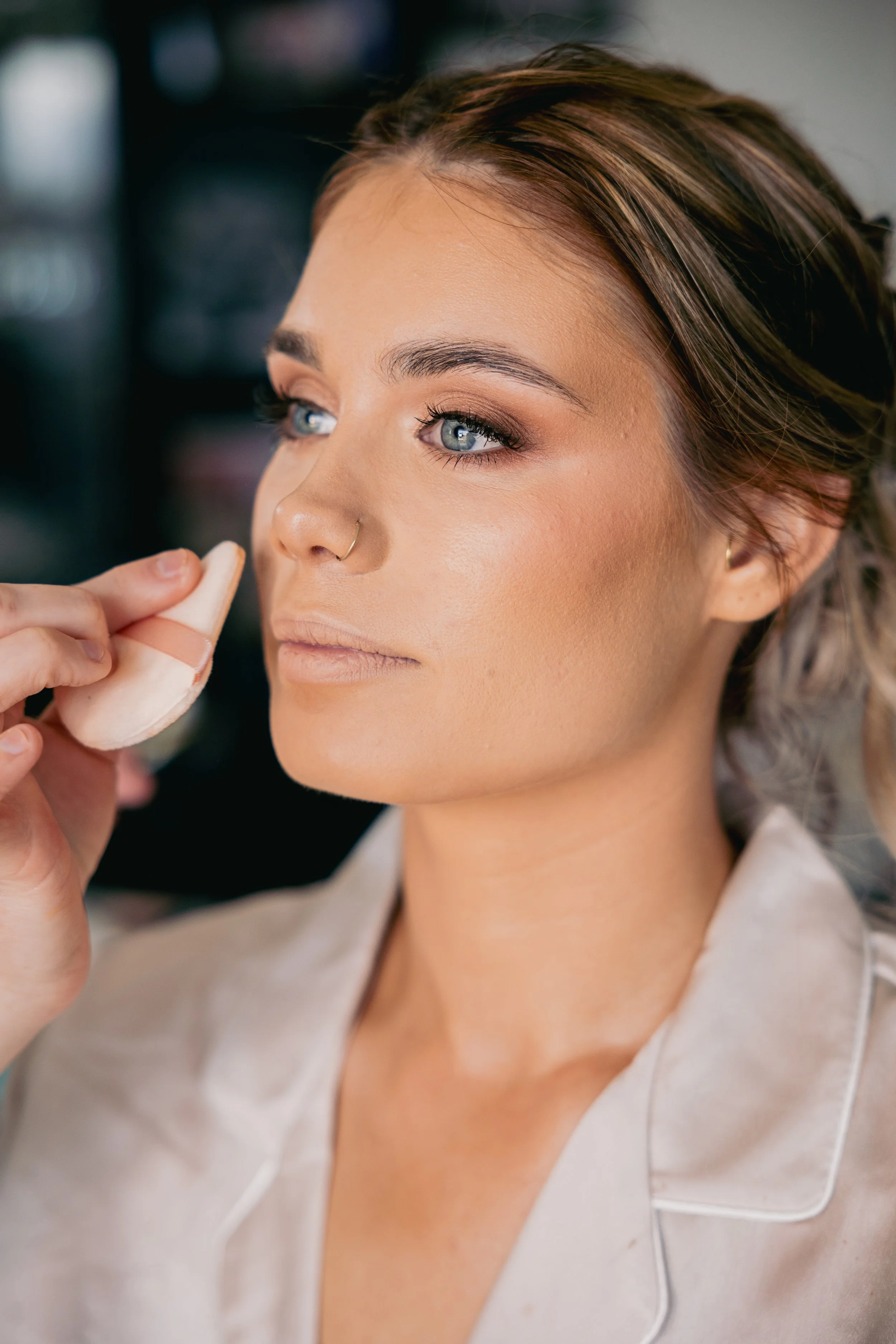 Woman with blue eyes and makeup having her face touched with a sponge by a makeup artist.