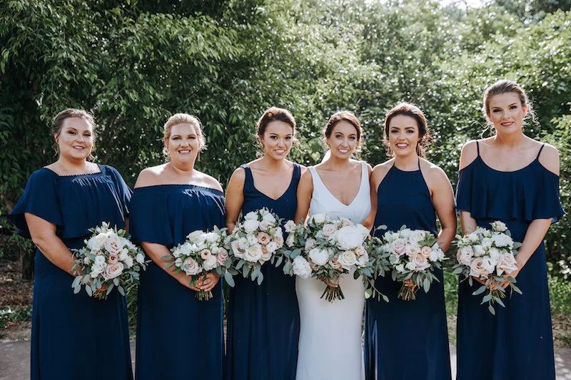 Six women in dresses standing outdoors, holding bouquets, at a wedding.