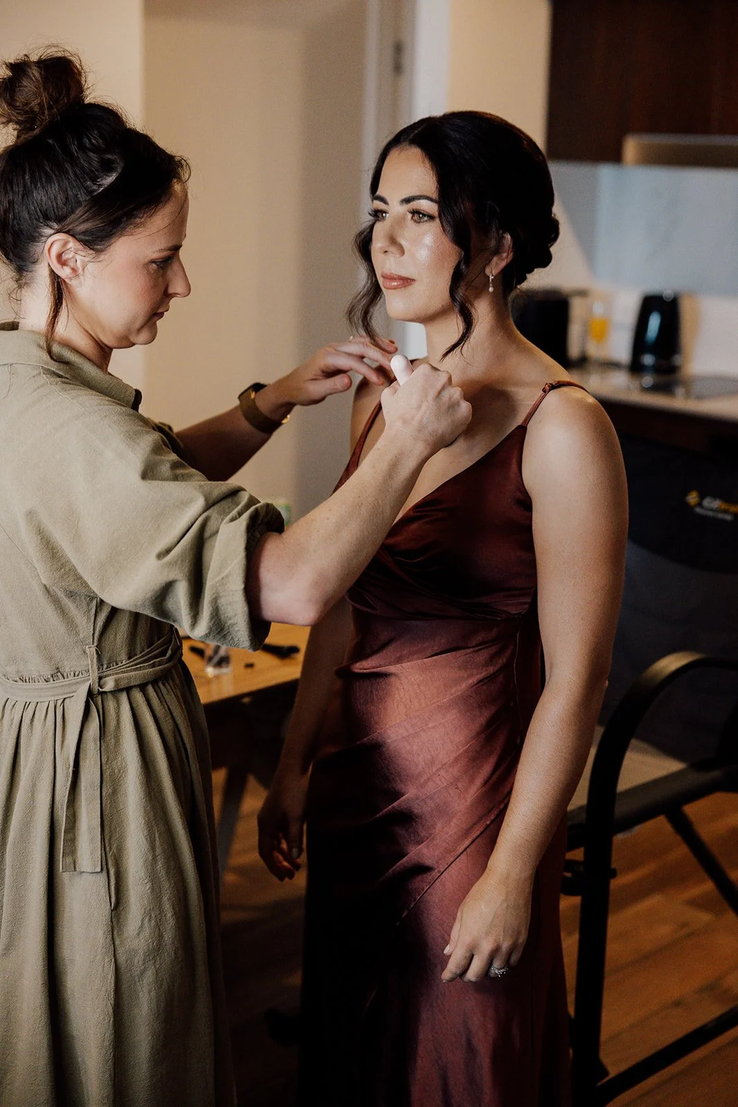 A woman in a satin burgundy dress gets her makeup done by a makeup artist in a beige dress.