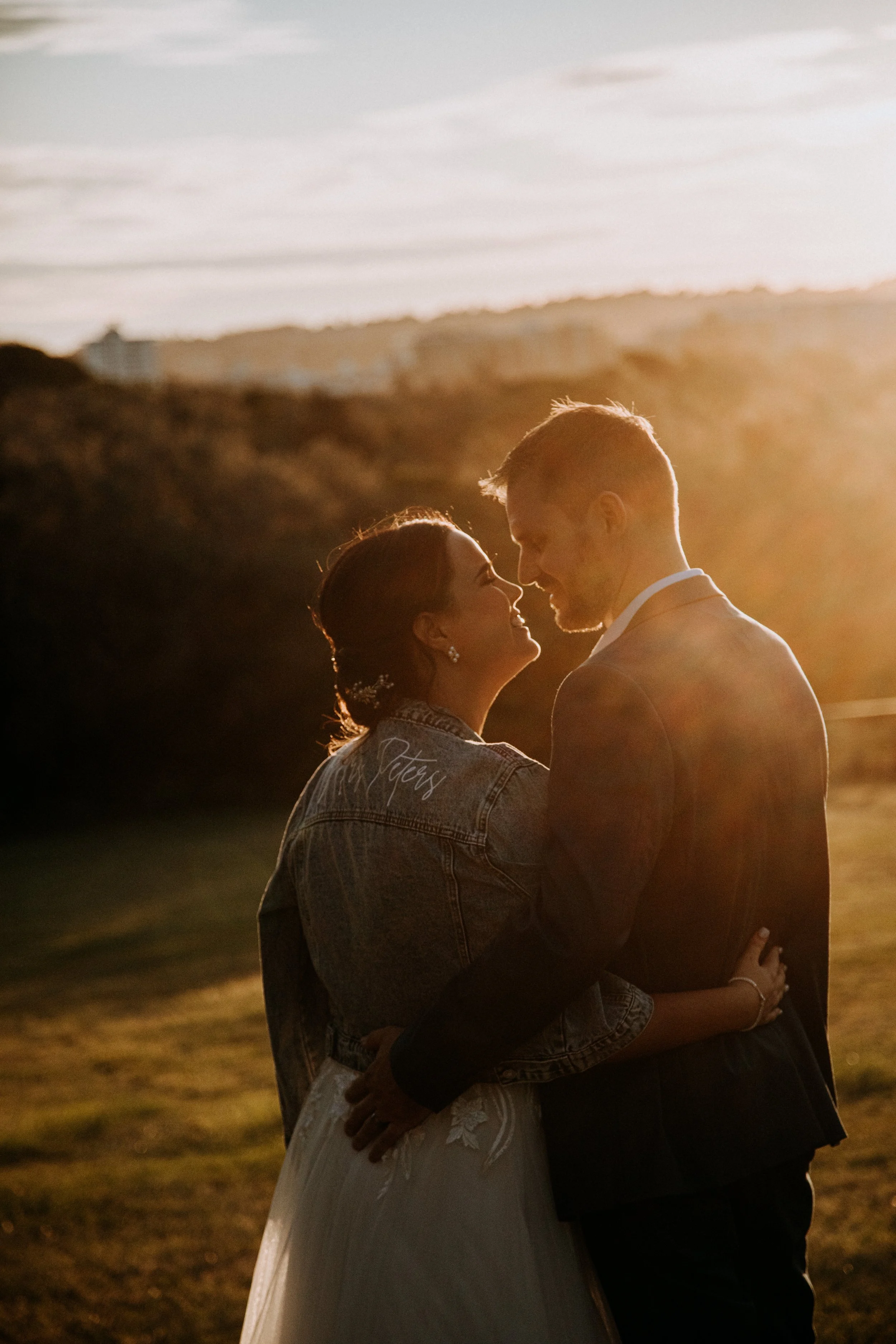 A bride and groom embrace outdoors during sunset, with the bride wearing a denim jacket and a white wedding dress, and the groom in a dark suit, standing close and smiling at each other.