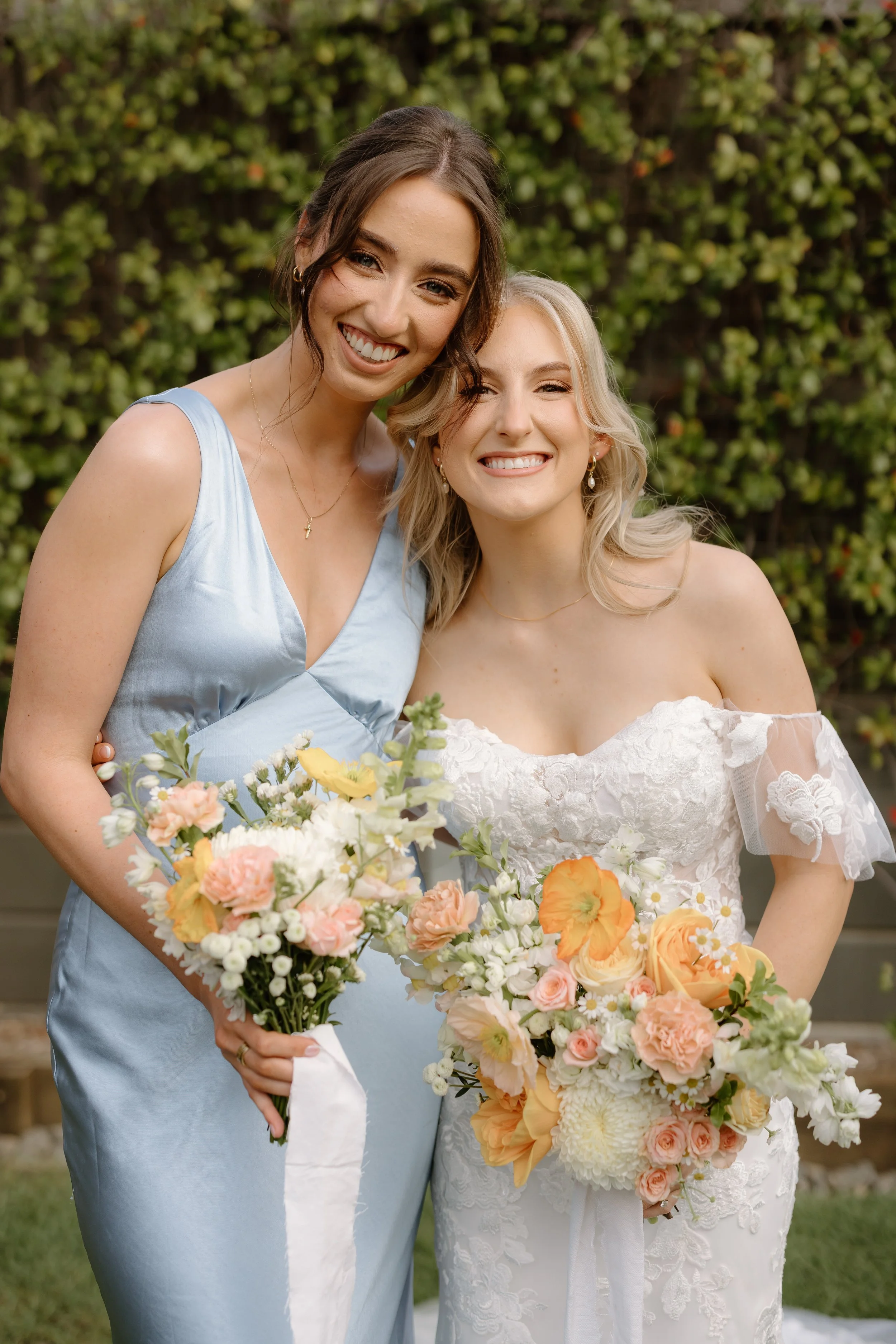 Two women smiling, holding bouquets, standing outdoors with a background of green foliage; one in a light blue dress and the other in a white lace wedding gown.